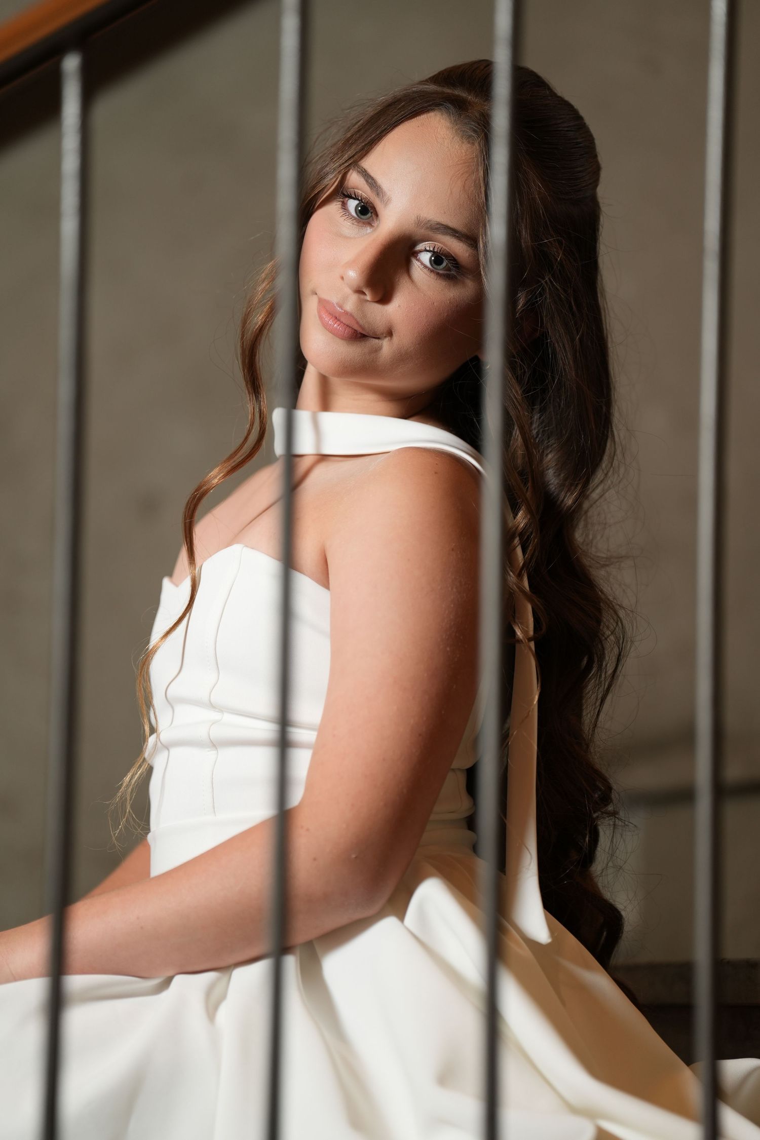 Young woman in white bridal gown with long wavy hair poses elegantly on staircase, gazing softly at camera.