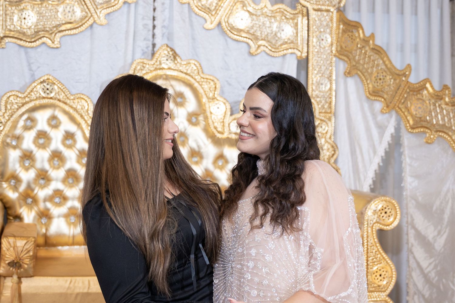 Two people laughing together in front of an ornate gold and white backdrop at a formal event.