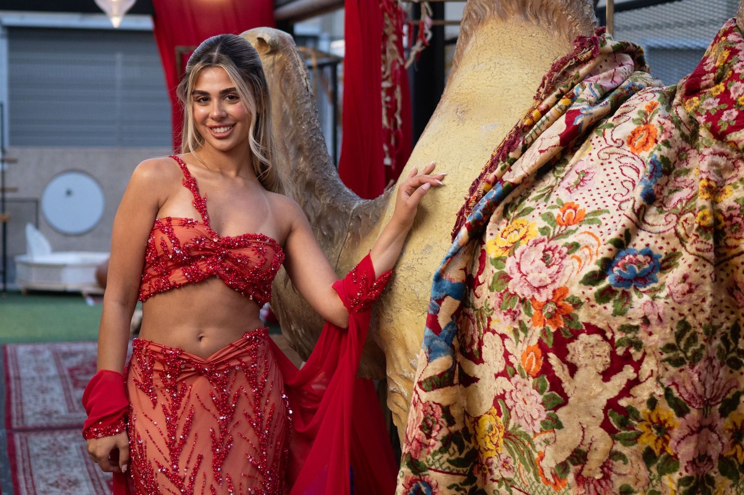 Woman in red embroidered crop top and skirt smiles beside ornate floral embroidered garment at outdoor market.