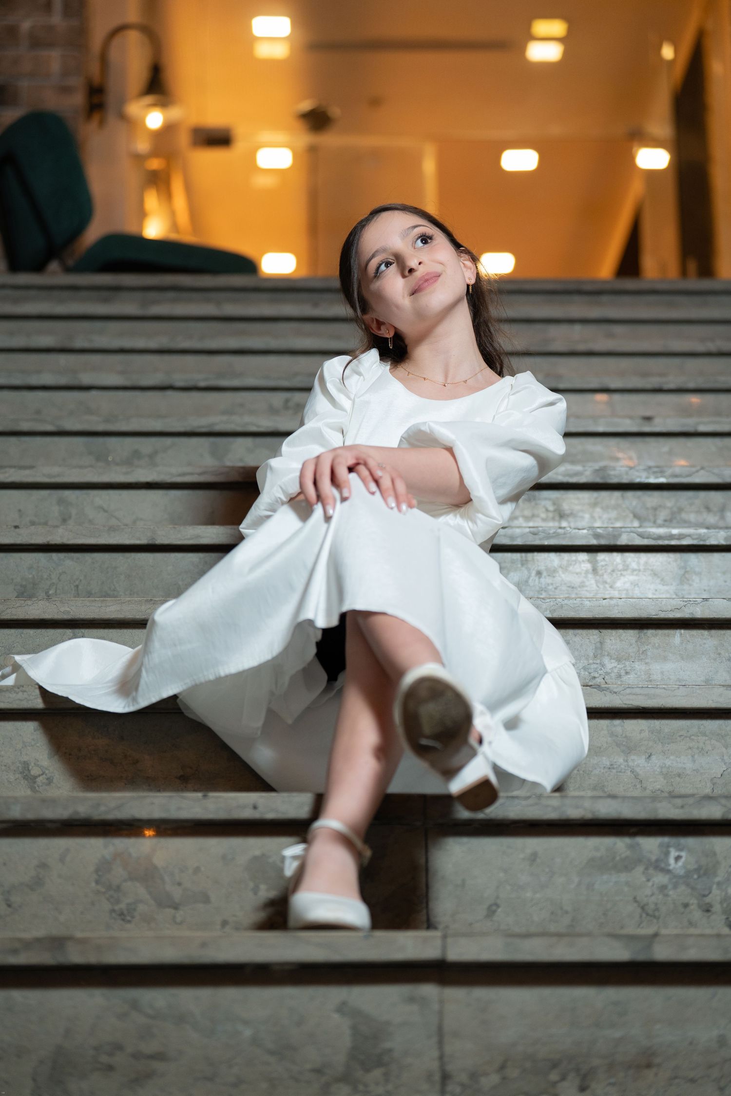 A figure in a flowing white dress reclines gracefully on wooden bleacher steps under warm indoor lighting.