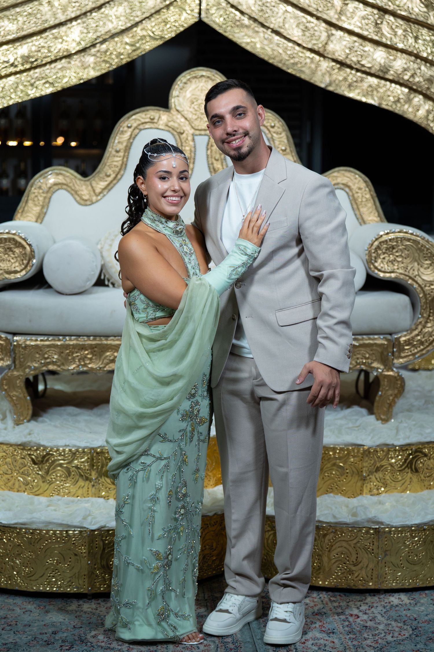 Elegant couple posing in formal attire before ornate gold throne, woman in mint green gown, man in beige suit.