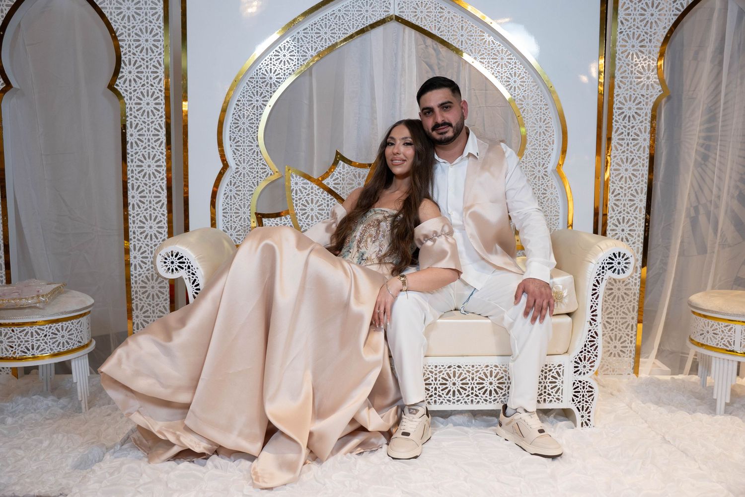 Wedding couple poses in elegant gold and white ballroom with ornate decor and flowing champagne gown.