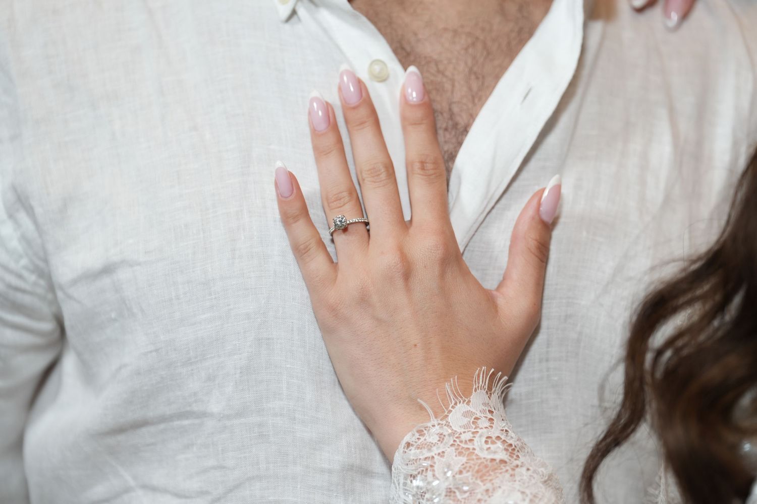 Close-up of a woman's hand with pink almond nails and a diamond engagement ring resting on a man's chest.