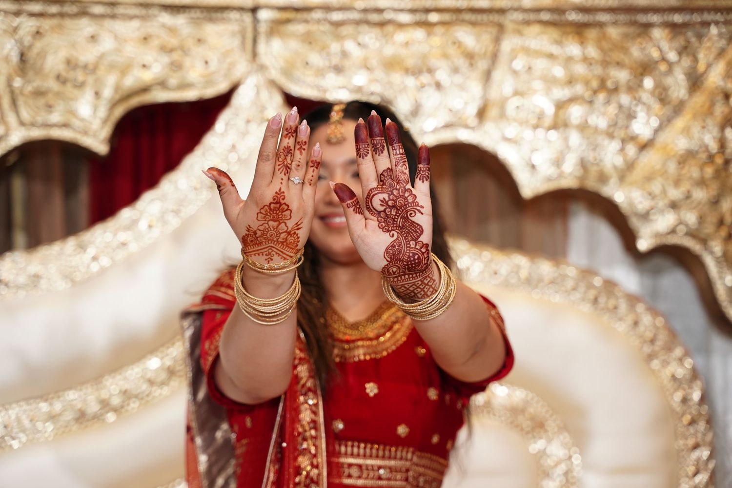 Indian bride in red lehenga with gold jewelry displays intricate henna mehndi designs on her hands at wedding ceremony.