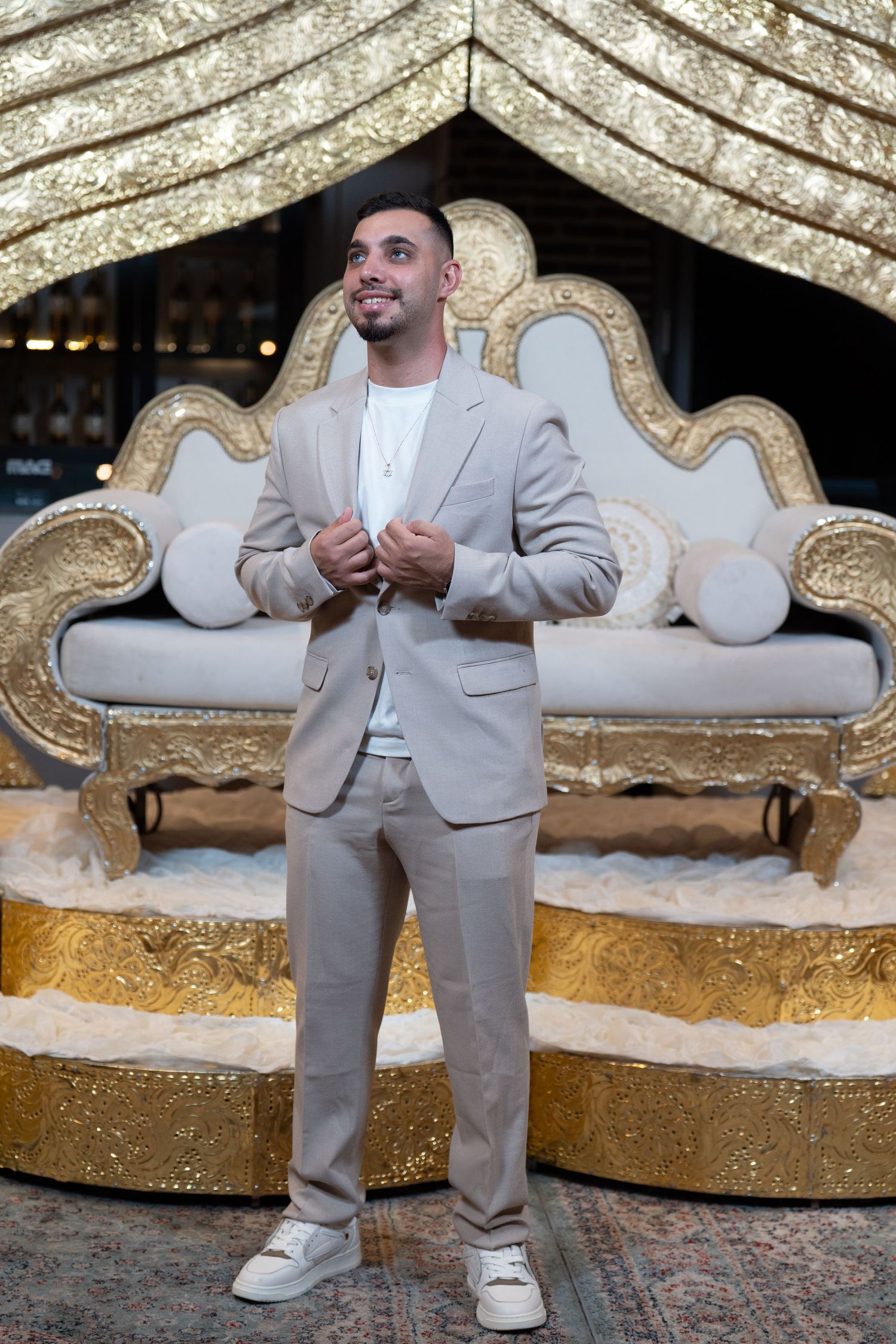 Man in beige suit stands before ornate gold and white throne sofa at elegant event venue.