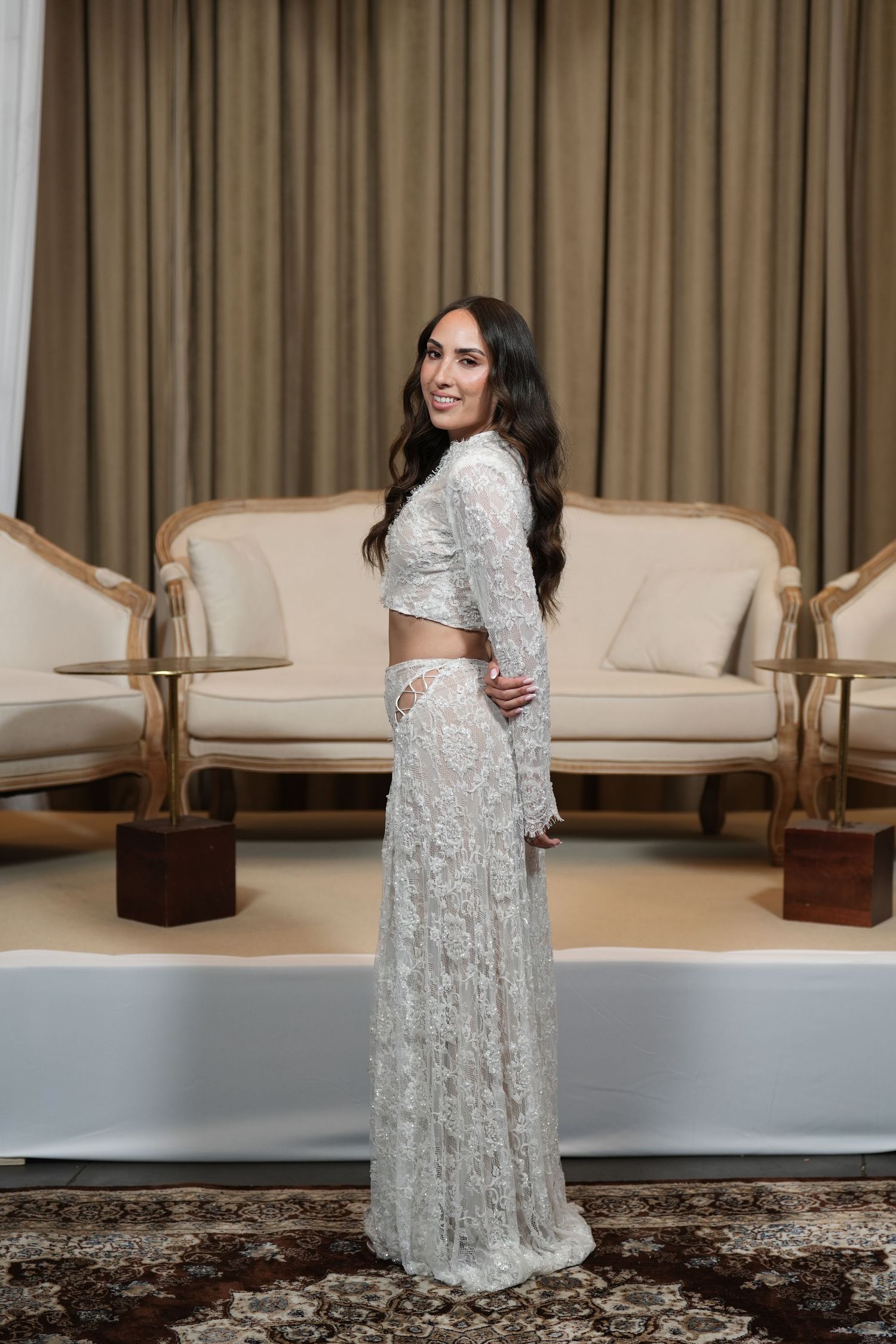 Woman in white lace two-piece bridal outfit posing elegantly in a luxurious room with beige furniture.