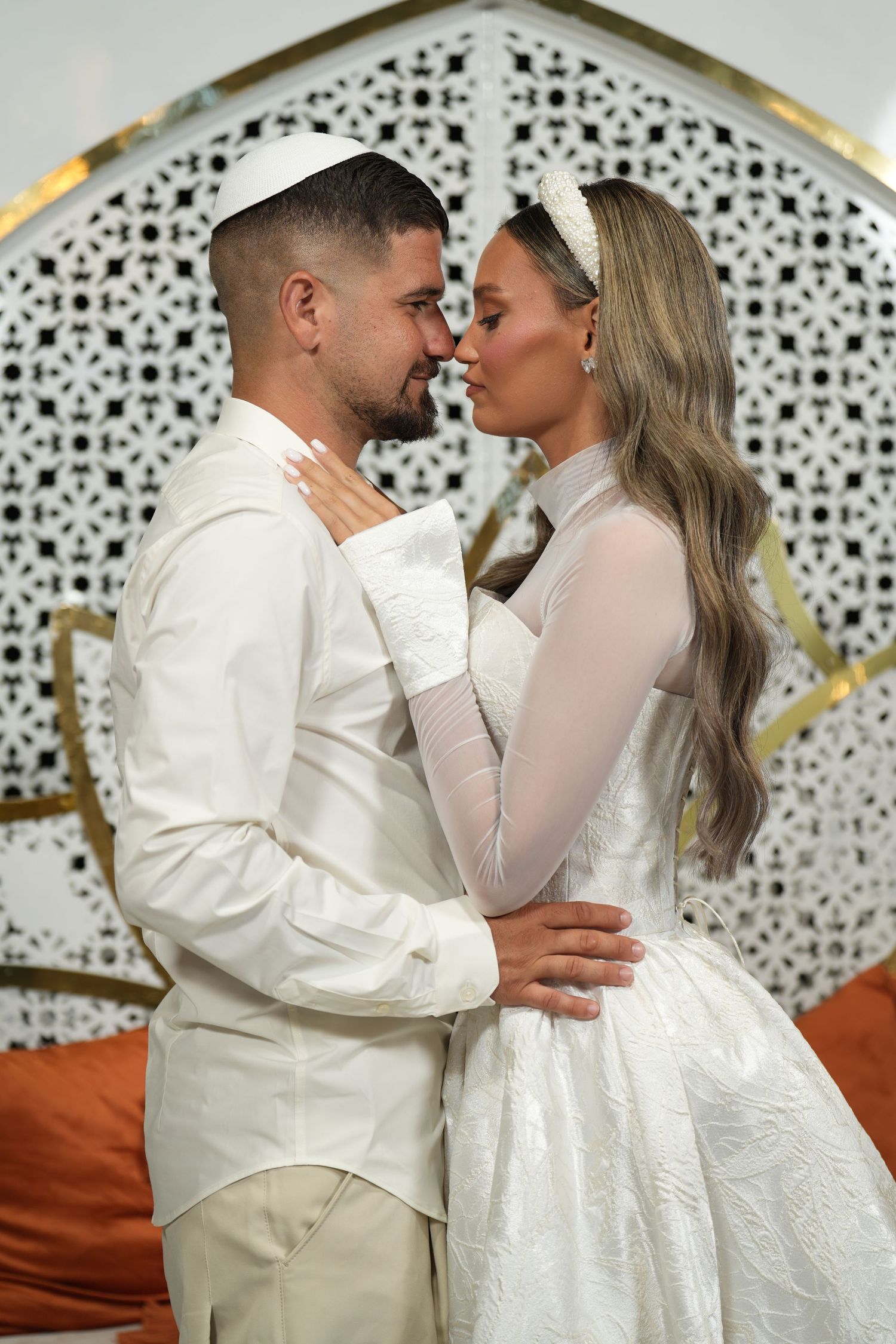 A couple in white wedding attire shares a romantic moment, the groom wearing a kippah and the bride in a lace gown.