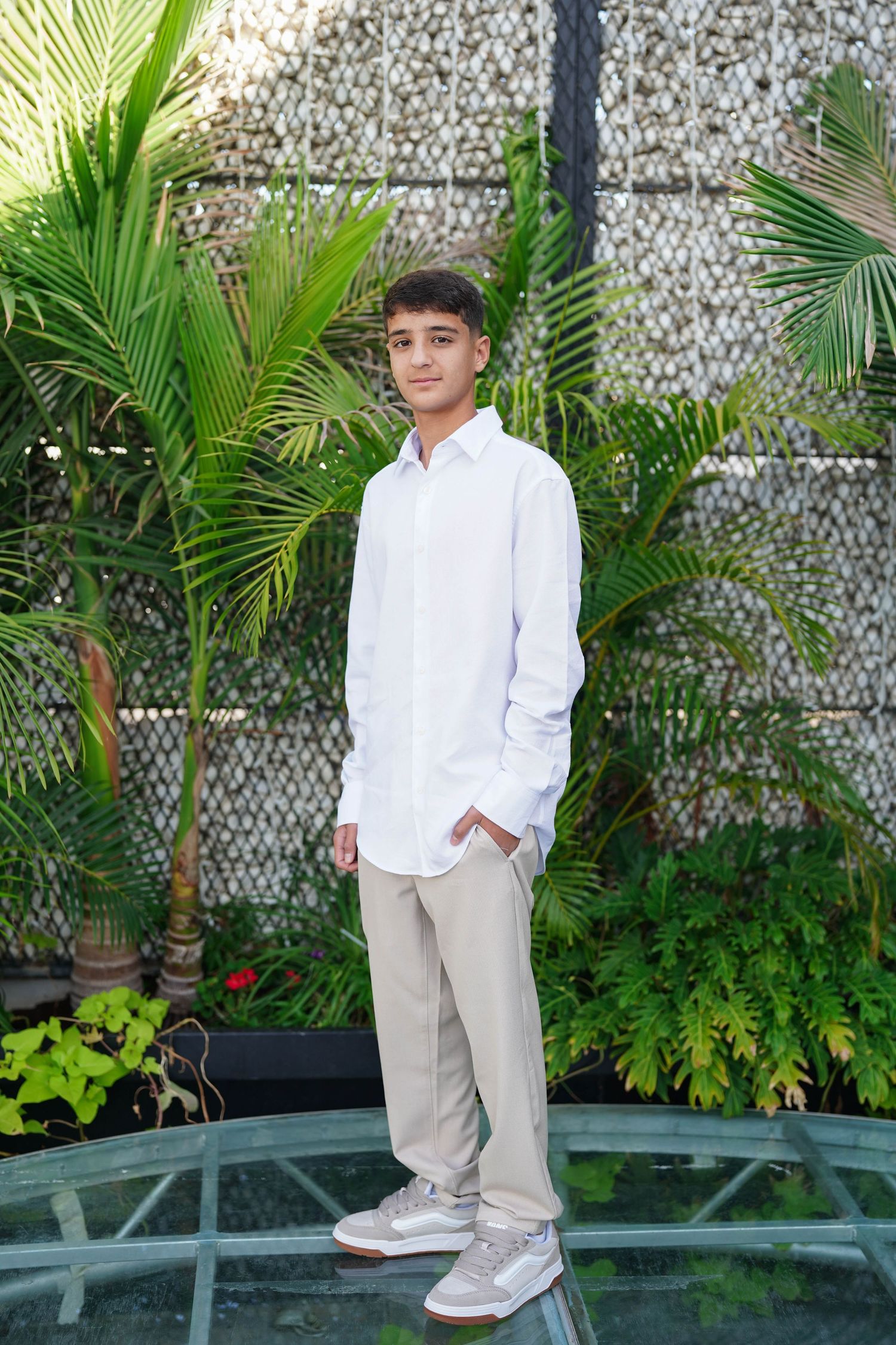 Child in formal white outfit and sneakers stands near palm trees and decorative wall.