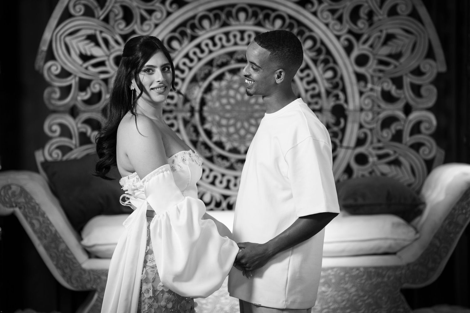 Black and white photo of a couple in elegant attire standing together in front of an ornate decorative backdrop.