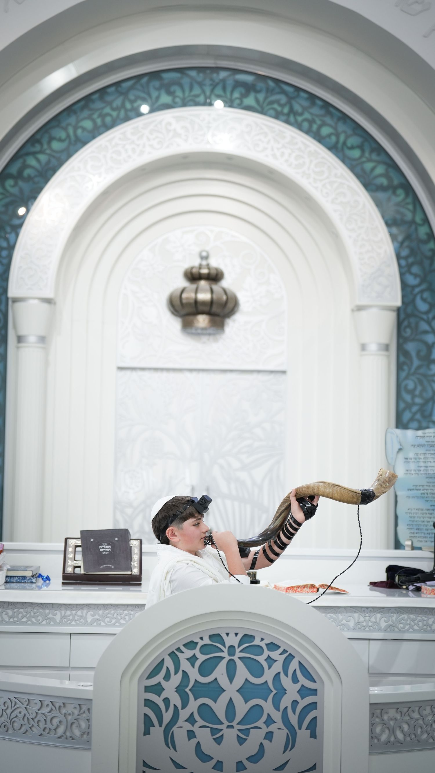 A young Jewish boy blows a shofar at a synagogue bimah, wearing tefillin, with ornate white and teal decor.