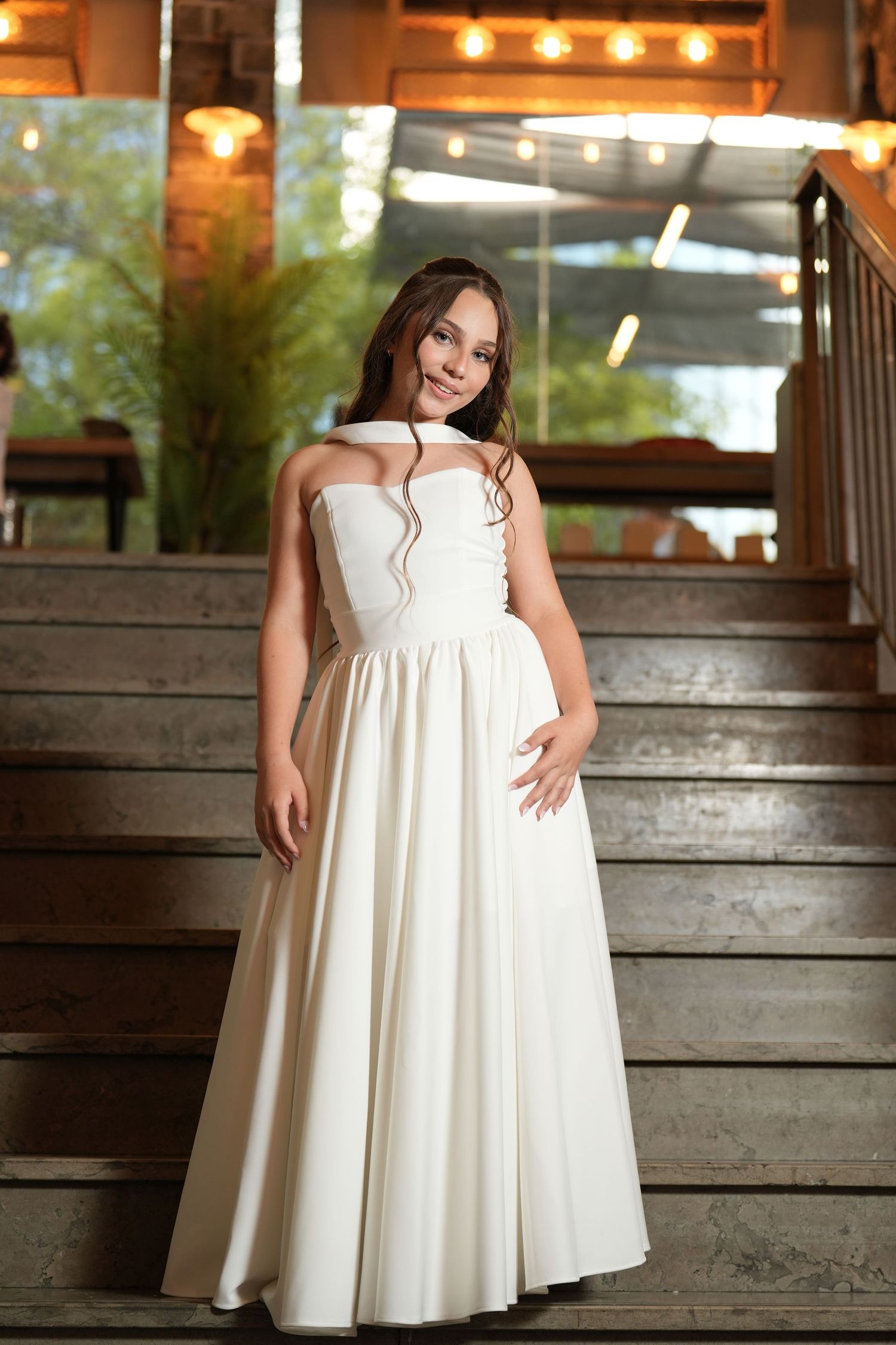 Young woman in elegant white strapless ball gown poses on wooden stairs with warm string lights in background.