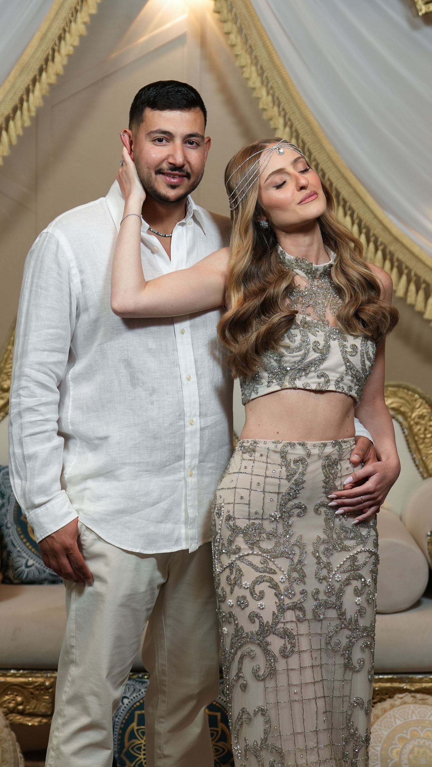 Couple posing together, man in white linen shirt, woman in ornate silver embroidered crop top and skirt with headpiece.