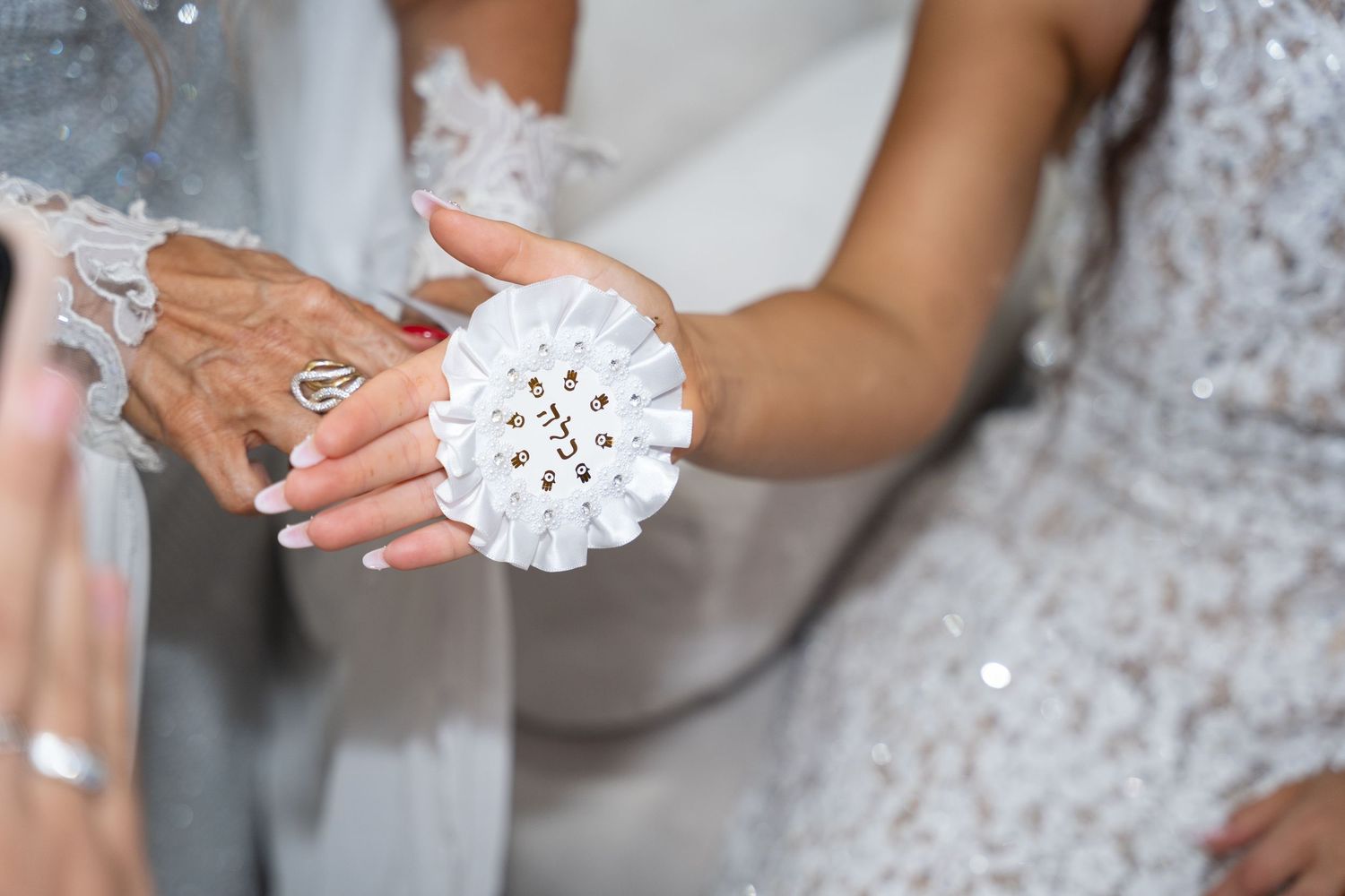 Close-up of bride wearing white floral wrist corsage with rhinestones, held by elderly woman with red nails at wedding.