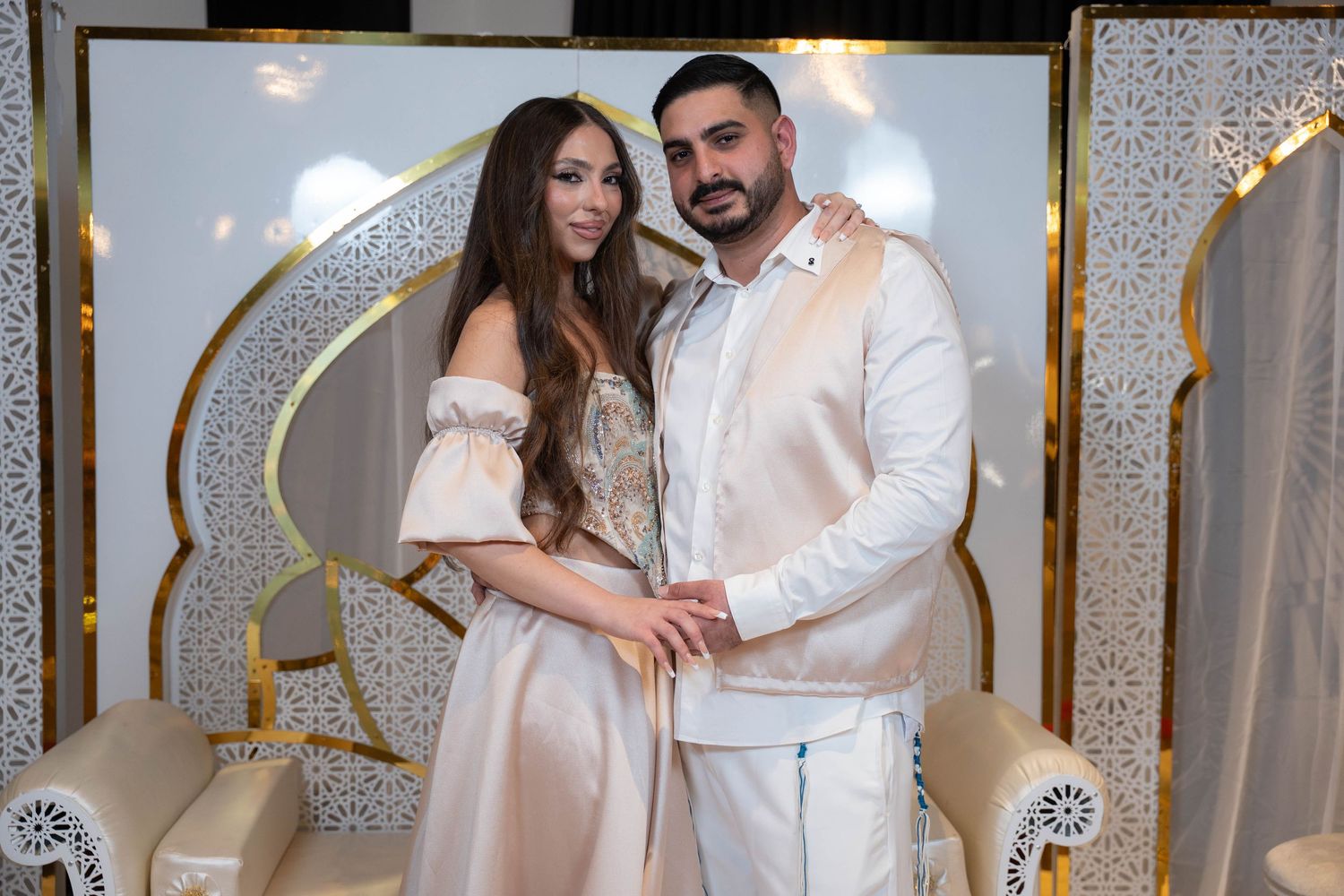 A couple poses together in elegant beige outfits against a decorative white and gold backdrop at a formal event.