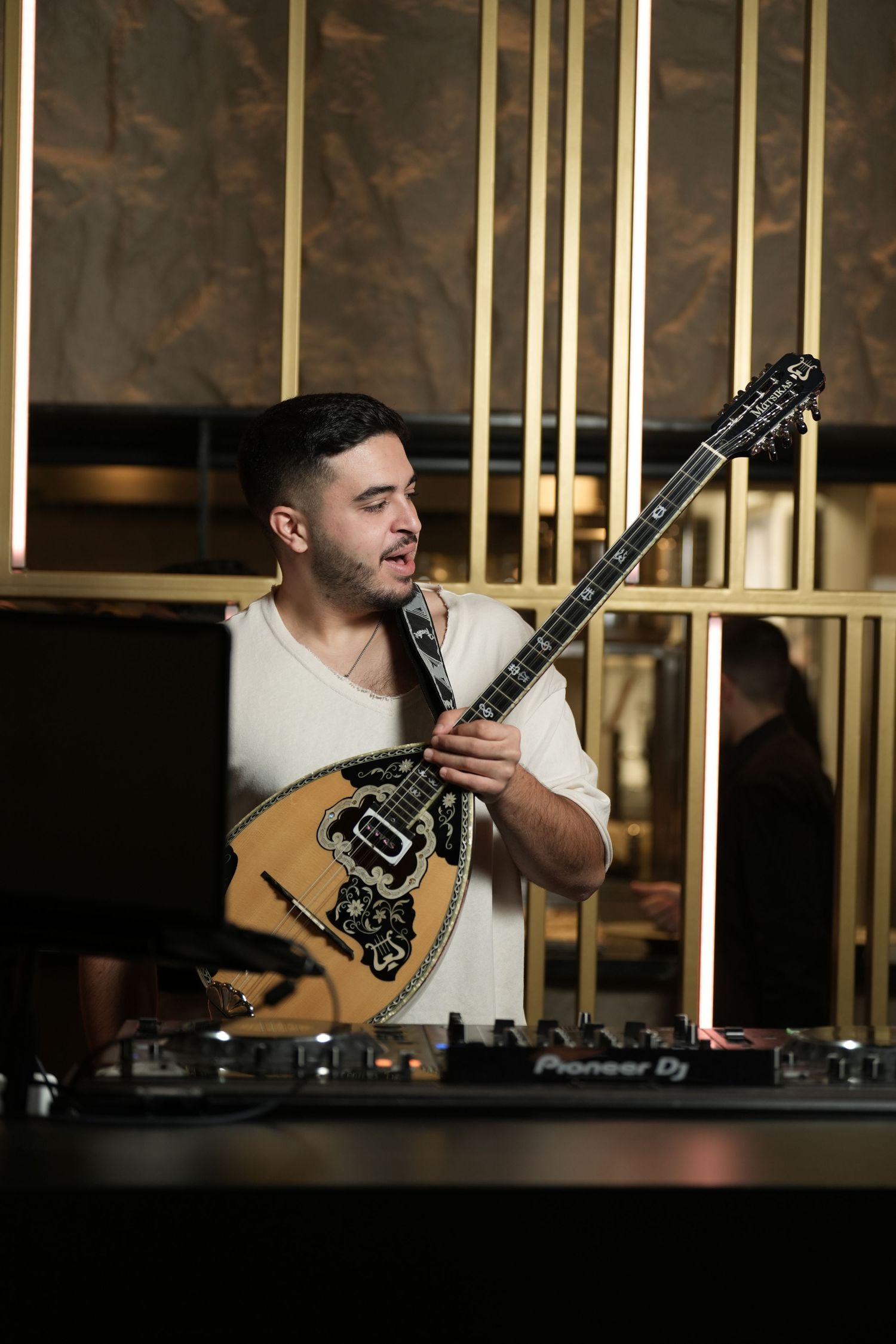 Musician performs on stage with a bouzouki instrument in a dimly lit venue with golden vertical bars in background.
