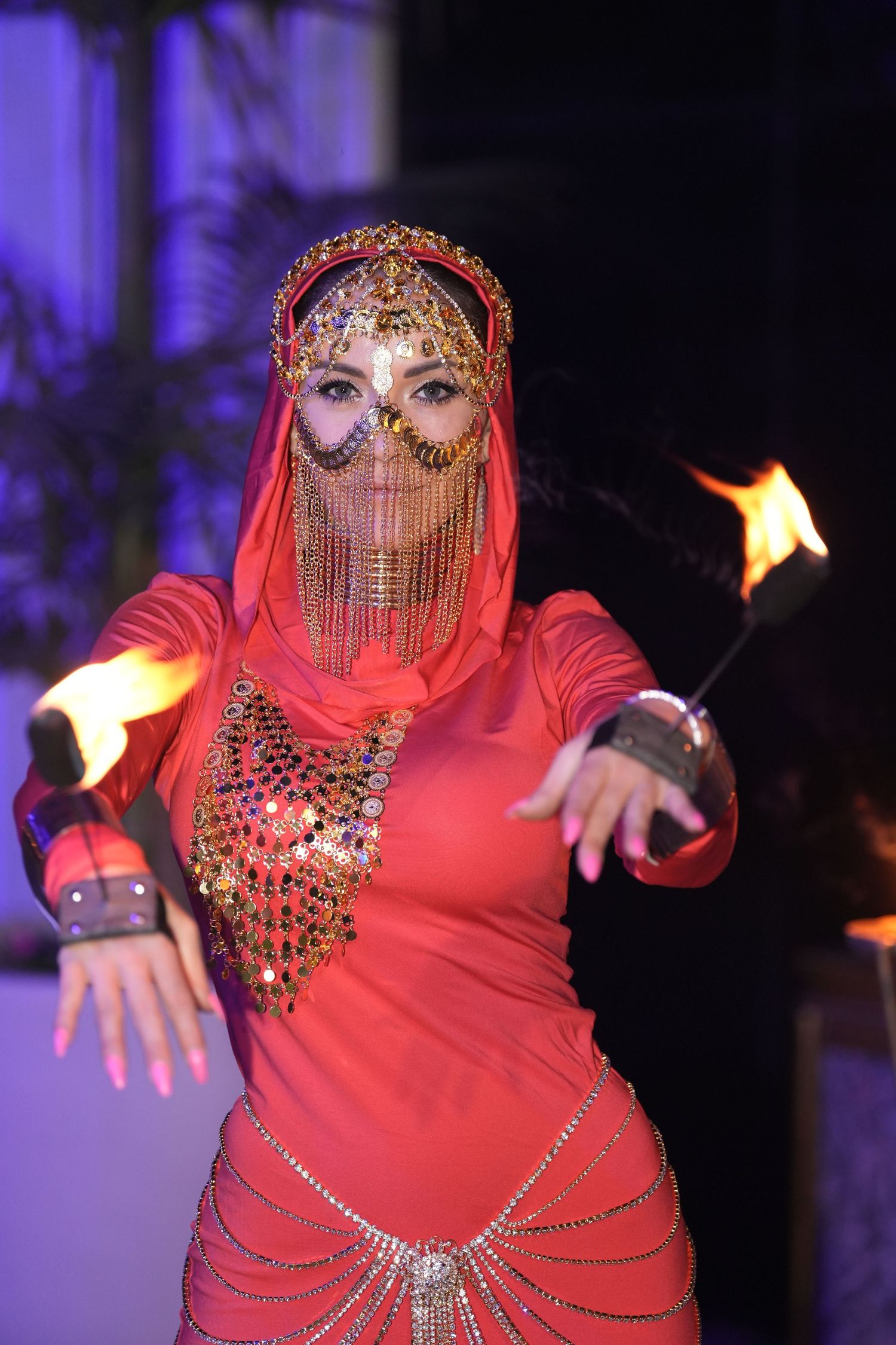 A fire dancer in red costume with jeweled veil and headdress performs with flames at an exotic cultural show.