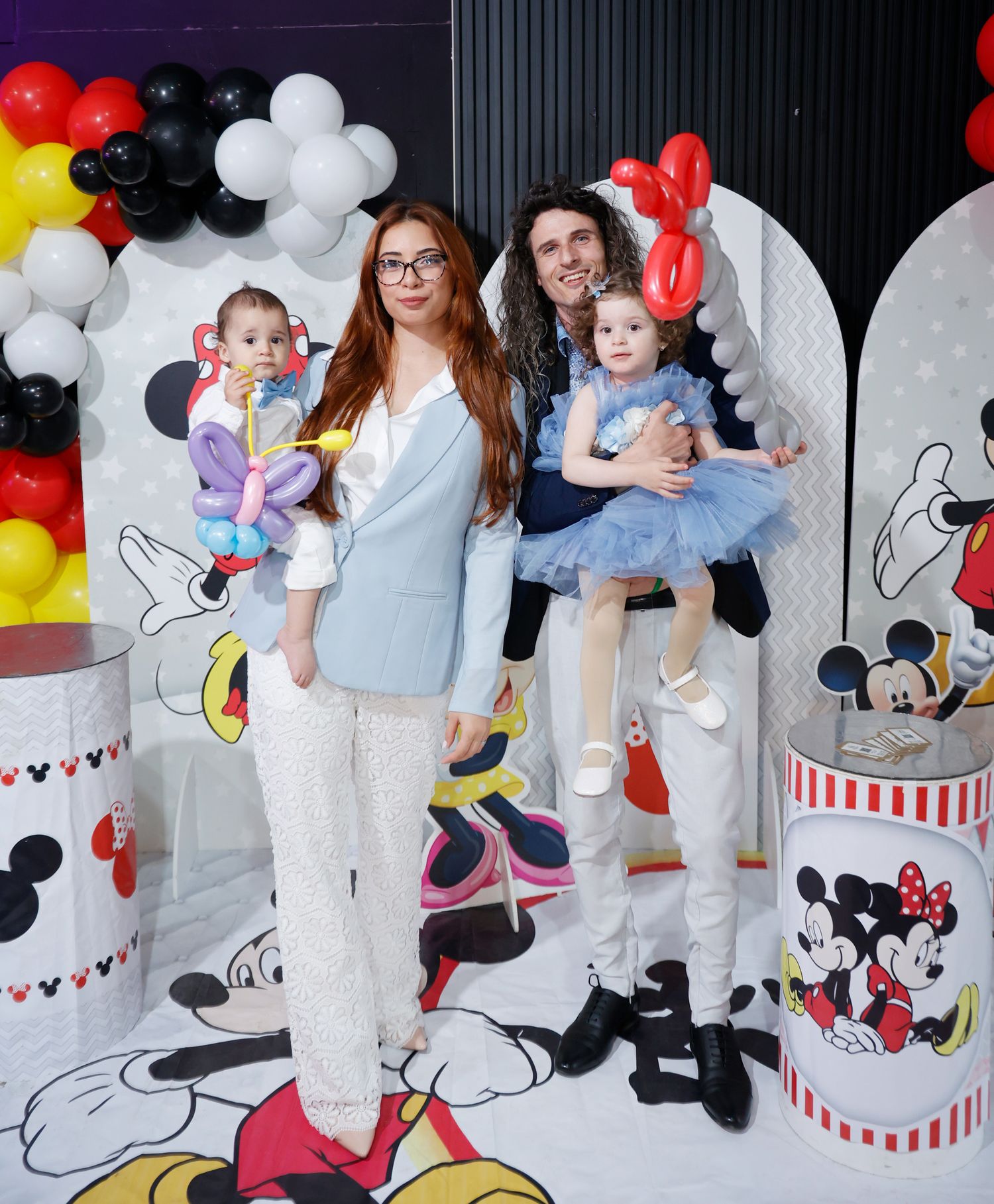 Family photo session against Mickey and Minnie Mouse themed backdrop with balloons and decorations.