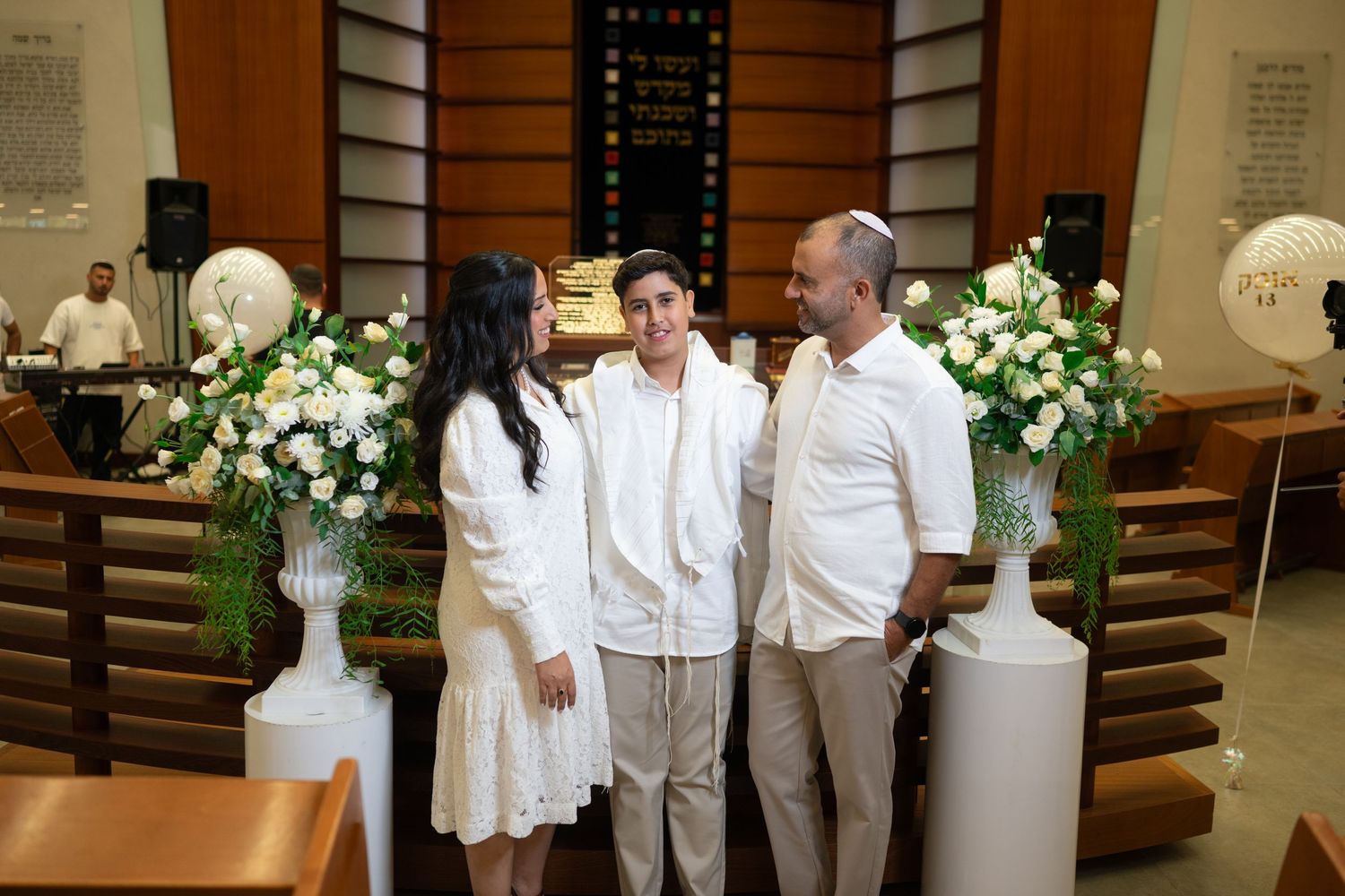 People in white clothing gathered around floral arrangements at an elegant indoor event.