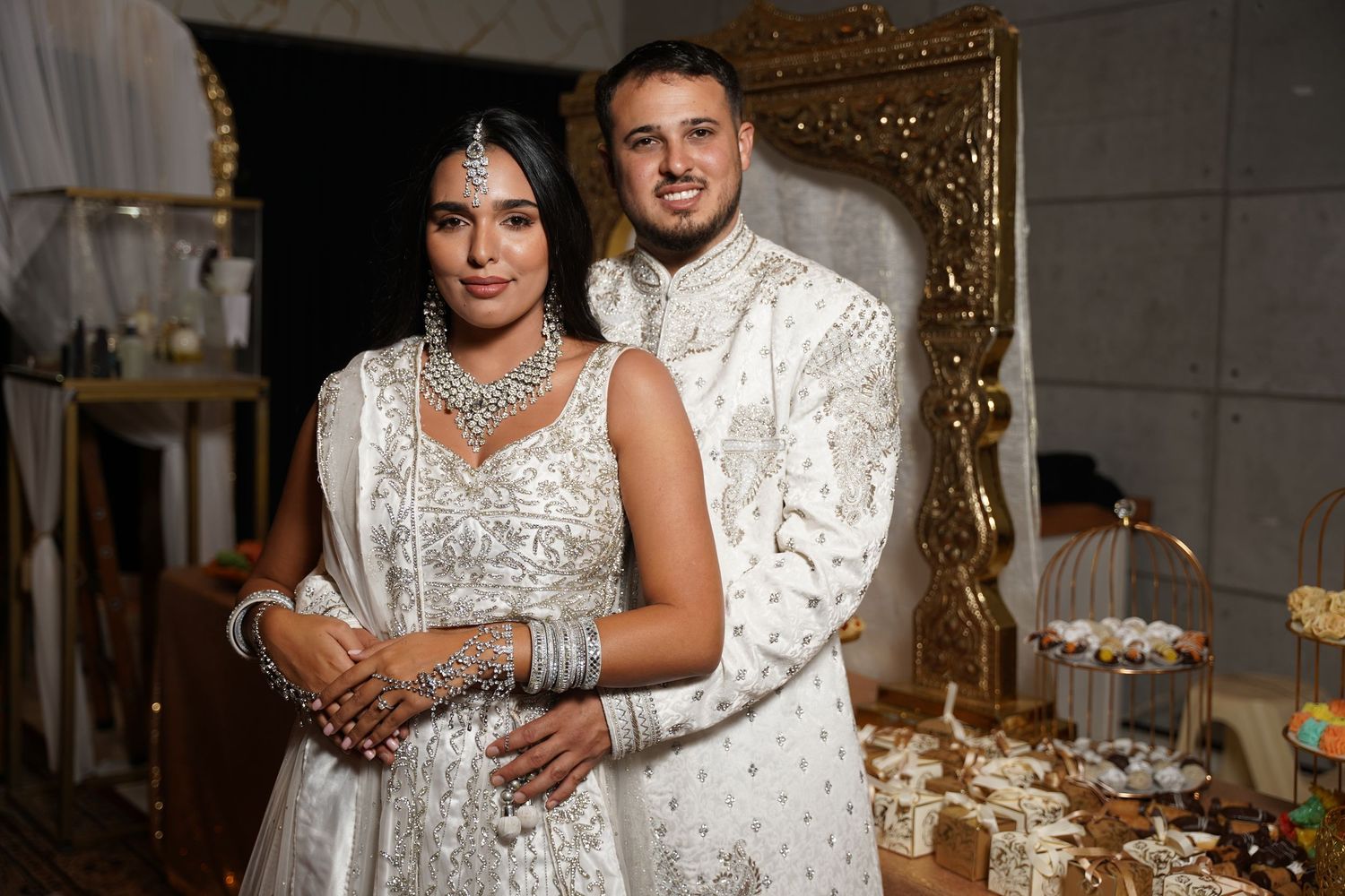 A couple in matching white traditional Indian wedding attire pose together against an ornate gold frame backdrop.