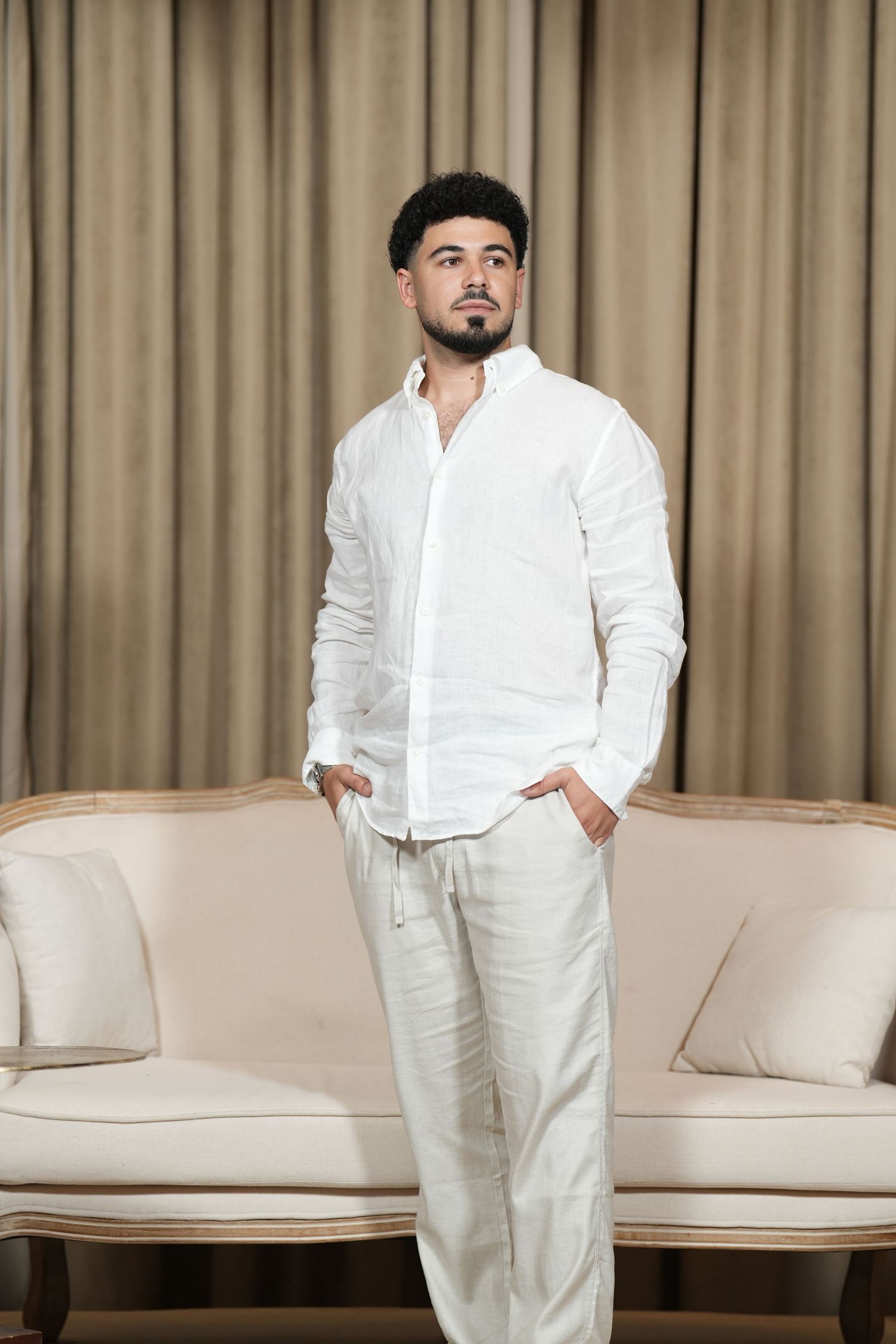 A young man in all-white linen shirt and pants stands confidently with hands in pockets before a beige sofa.