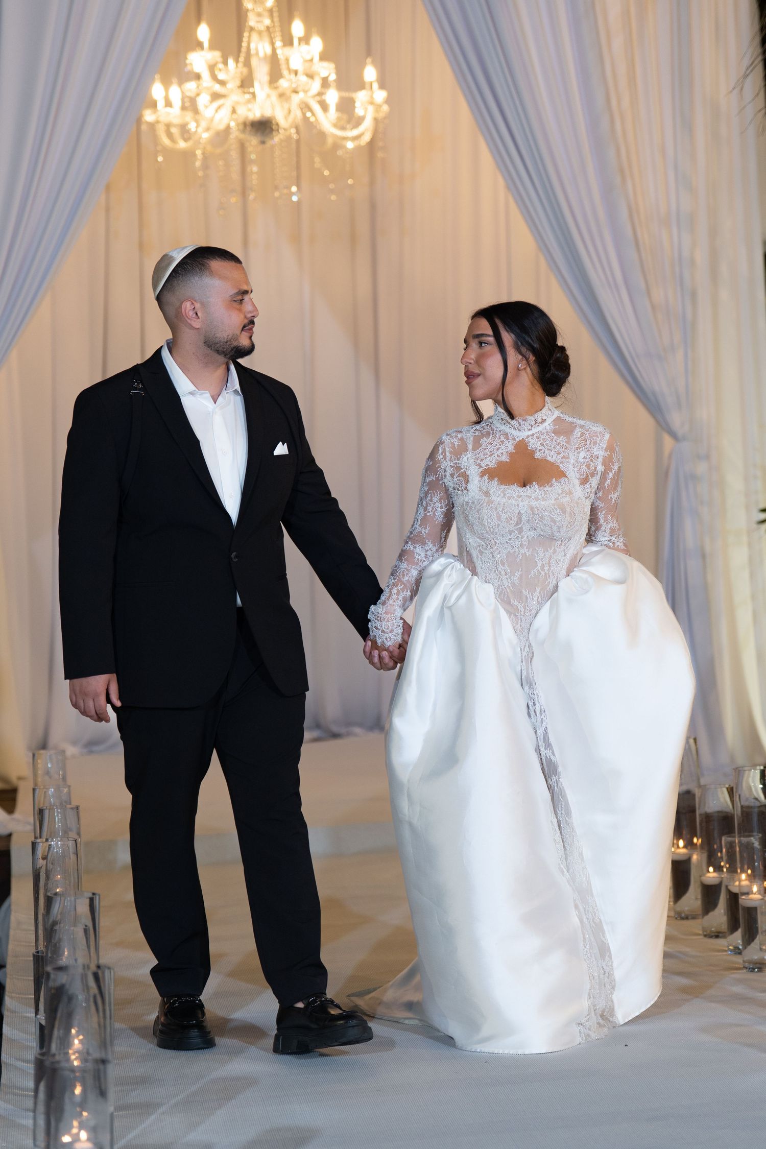 Bride in lace gown and groom in black suit hold hands at elegant Jewish wedding ceremony with chandelier.