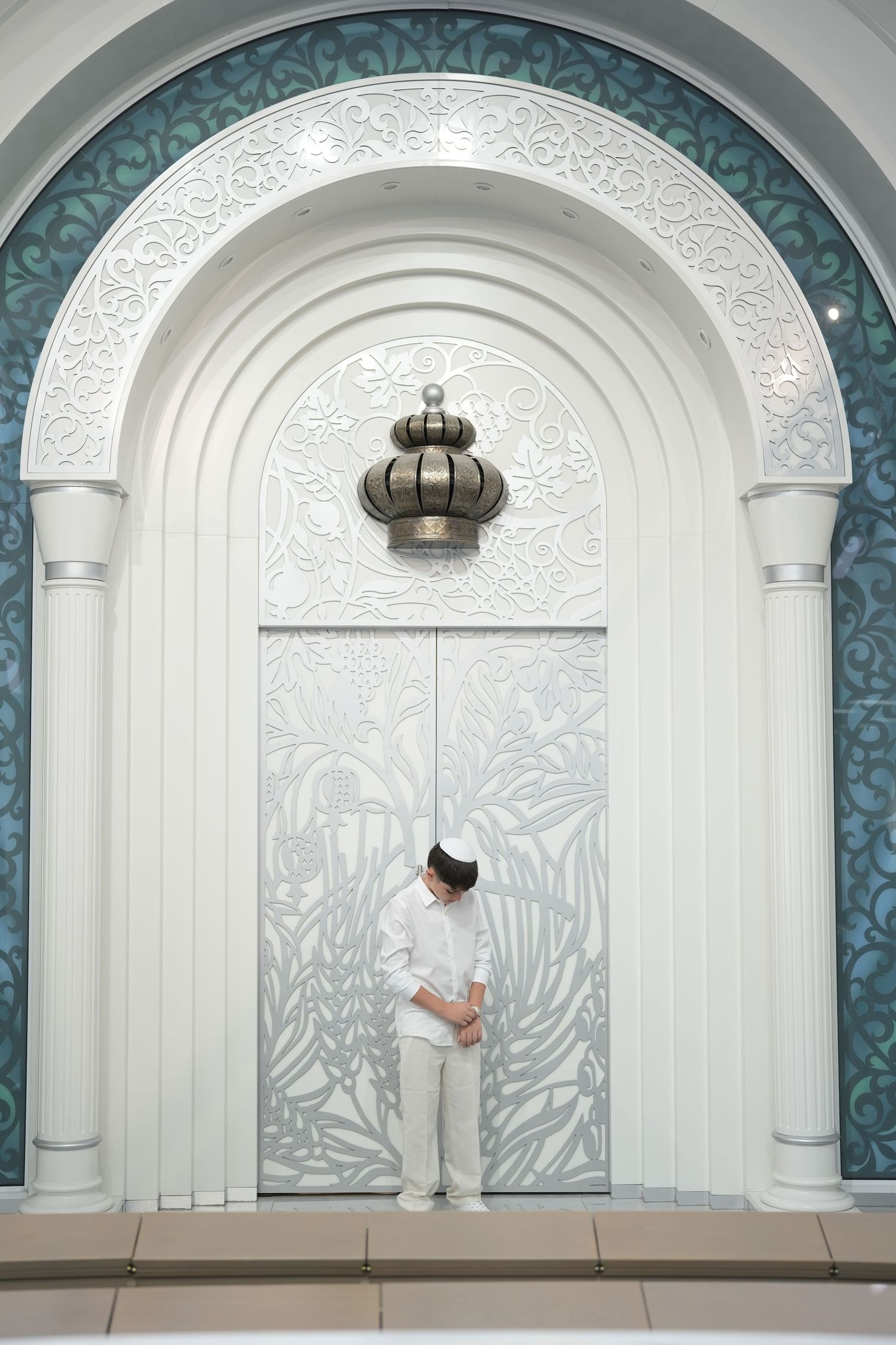 Person in white attire standing before an ornate white arched mosque door with floral carvings and a decorative lantern.