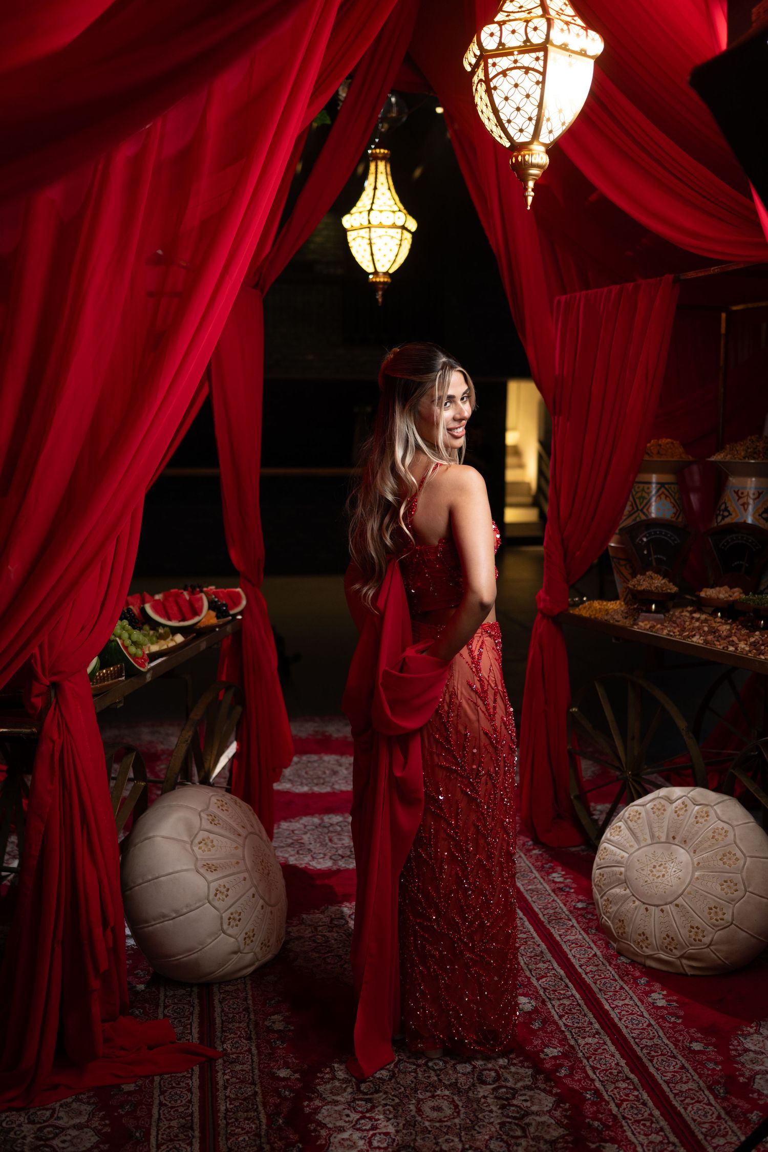 Woman in red beaded gown stands under Moroccan lanterns amid red drapes and ornate decor.