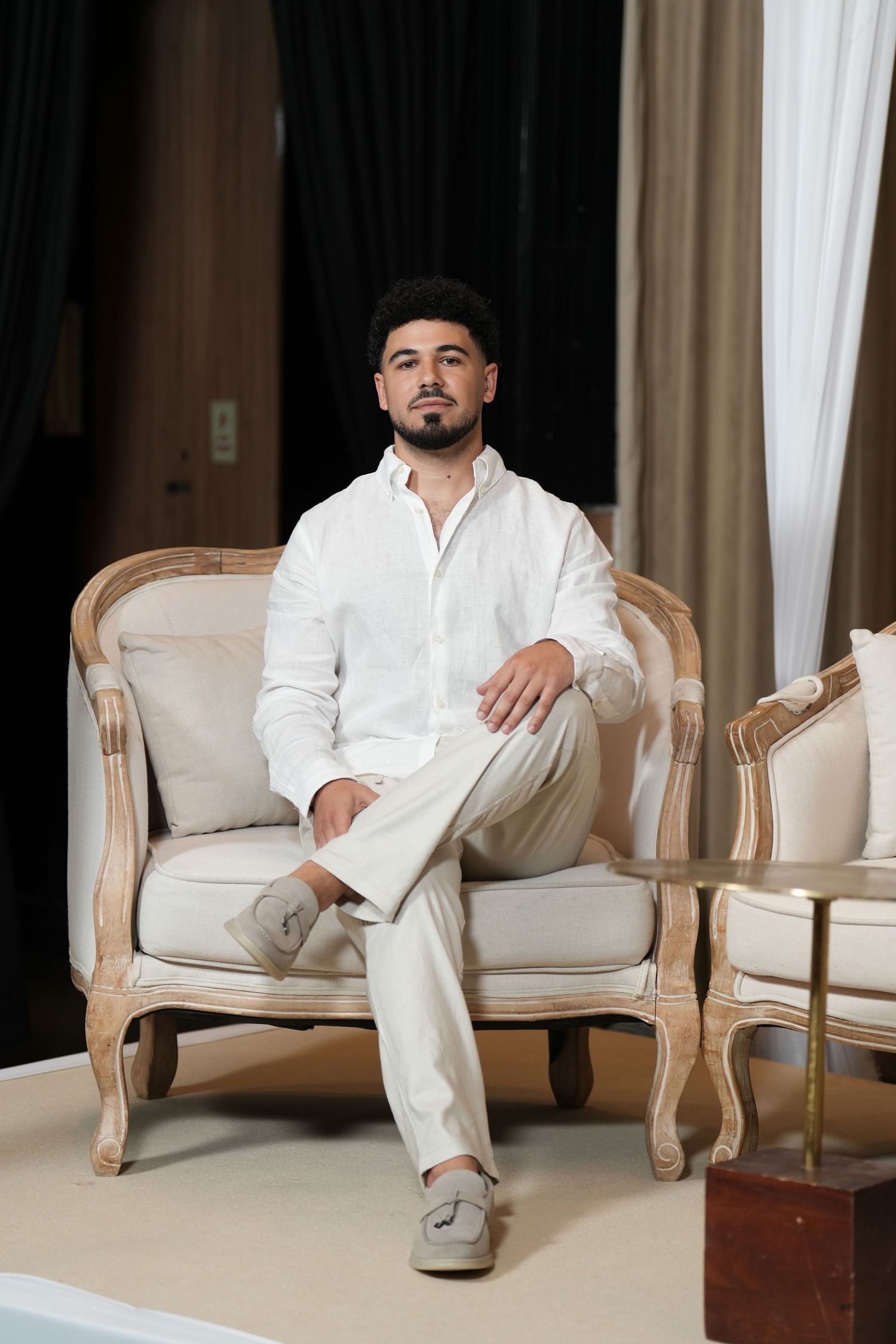 Young man in white linen shirt and beige pants sitting confidently on ornate wooden armchair in elegant room.