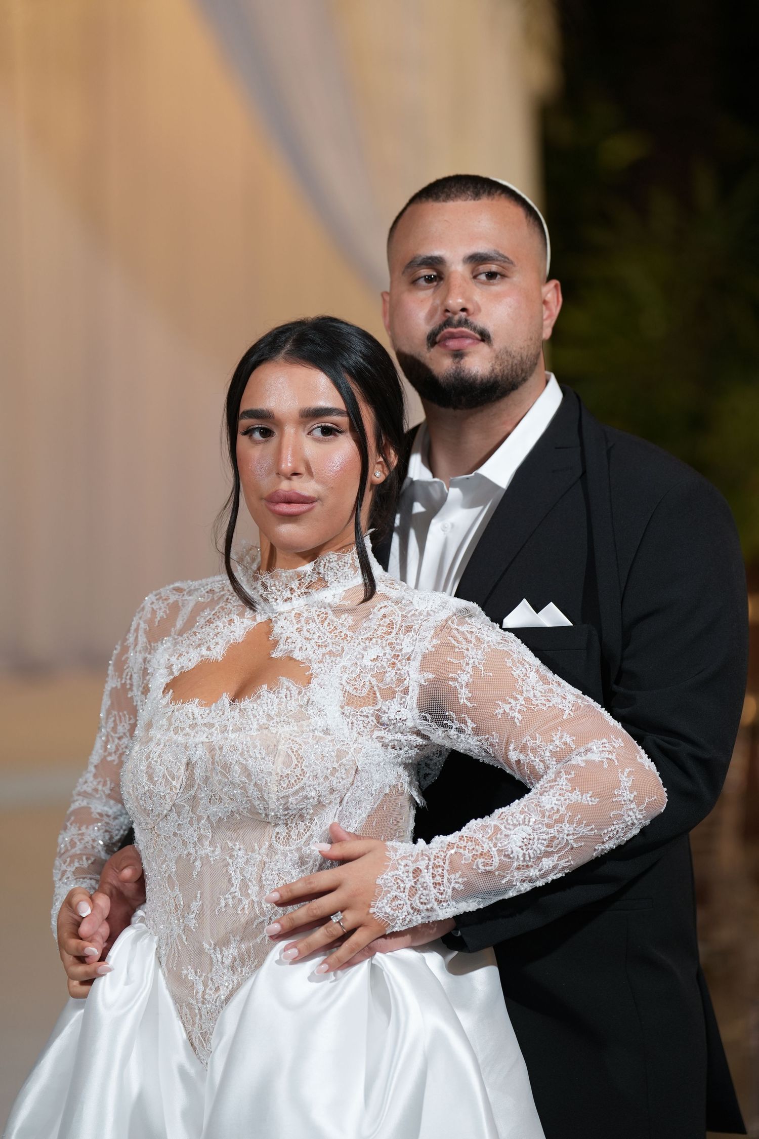 A bride in a lace long-sleeve white gown poses with a groom in a black tuxedo at their wedding ceremony.