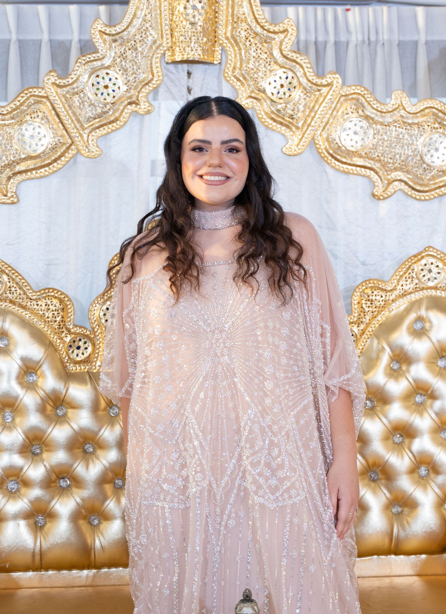 Person wearing a sparkly blush pink dress poses in front of an ornate gold throne backdrop.