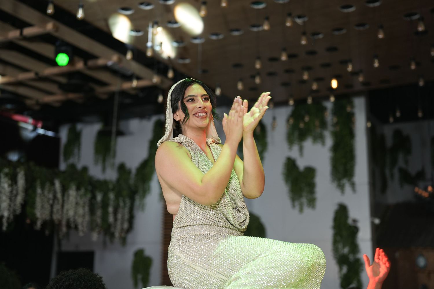 Woman in sparkly dress clapping and smiling on stage with green plant decorations.