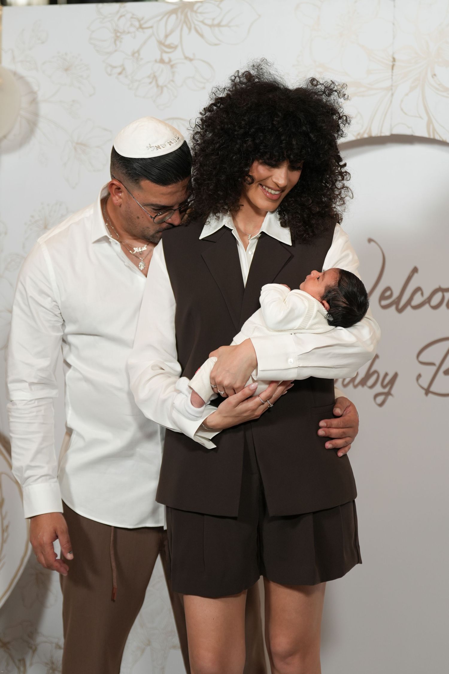 A couple in matching brown and white outfits smiles while holding a newborn baby at a welcome celebration.