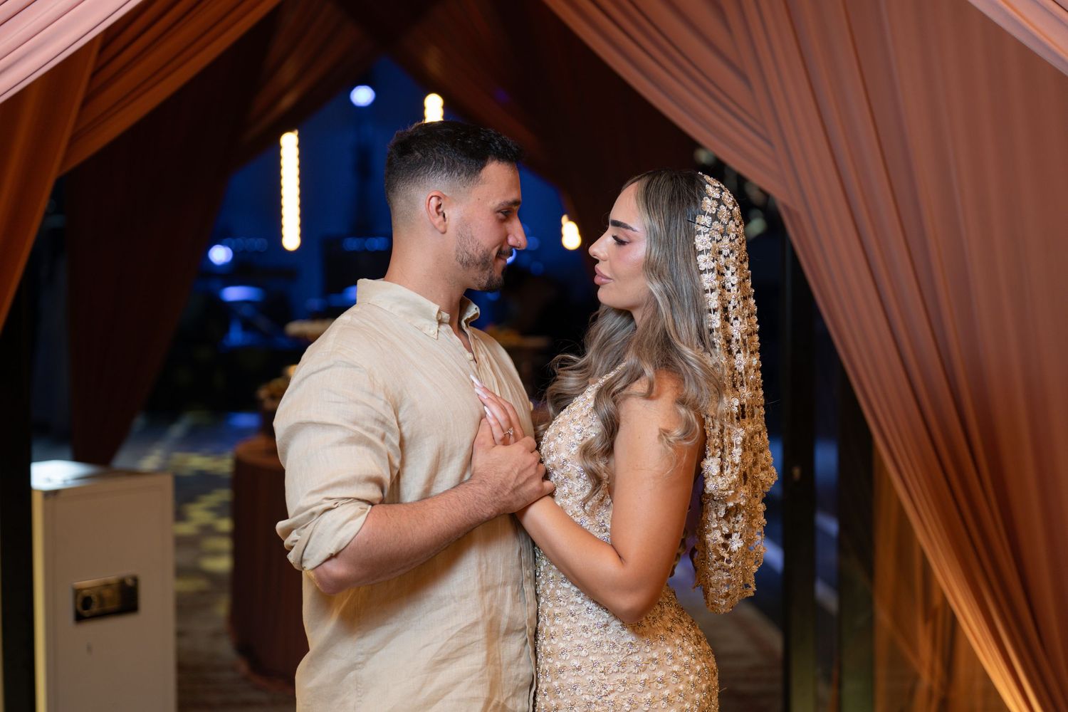Couple in elegant traditional attire sharing a romantic moment under a decorative arch at a wedding ceremony.