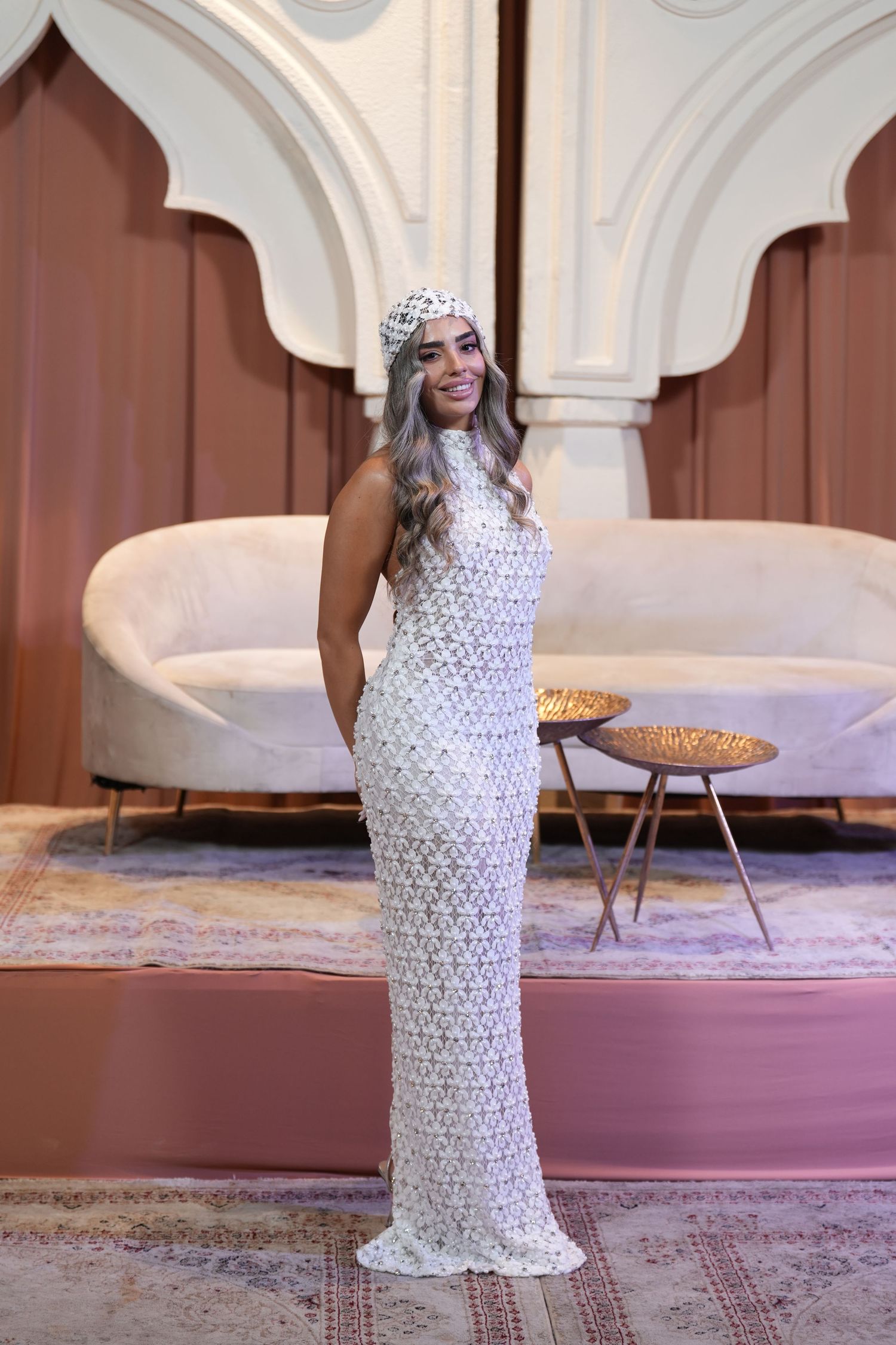 Woman in white lace gown and matching headpiece poses elegantly in a stylish room with arched decor.