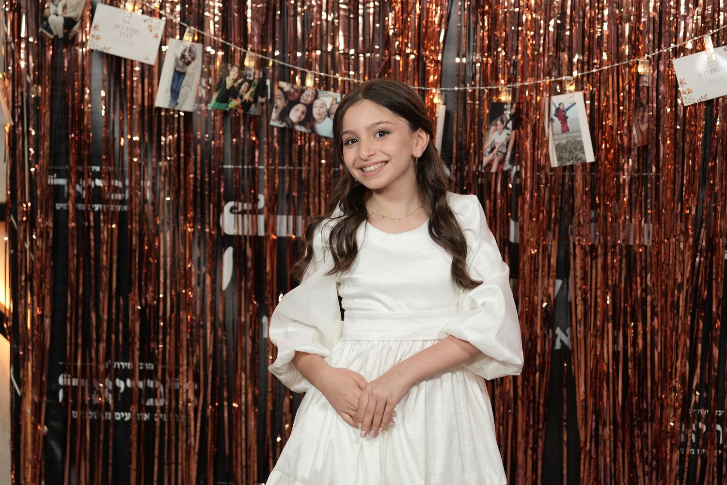 A person wearing a white dress poses against a metallic copper-colored tinsel backdrop at a party or event.