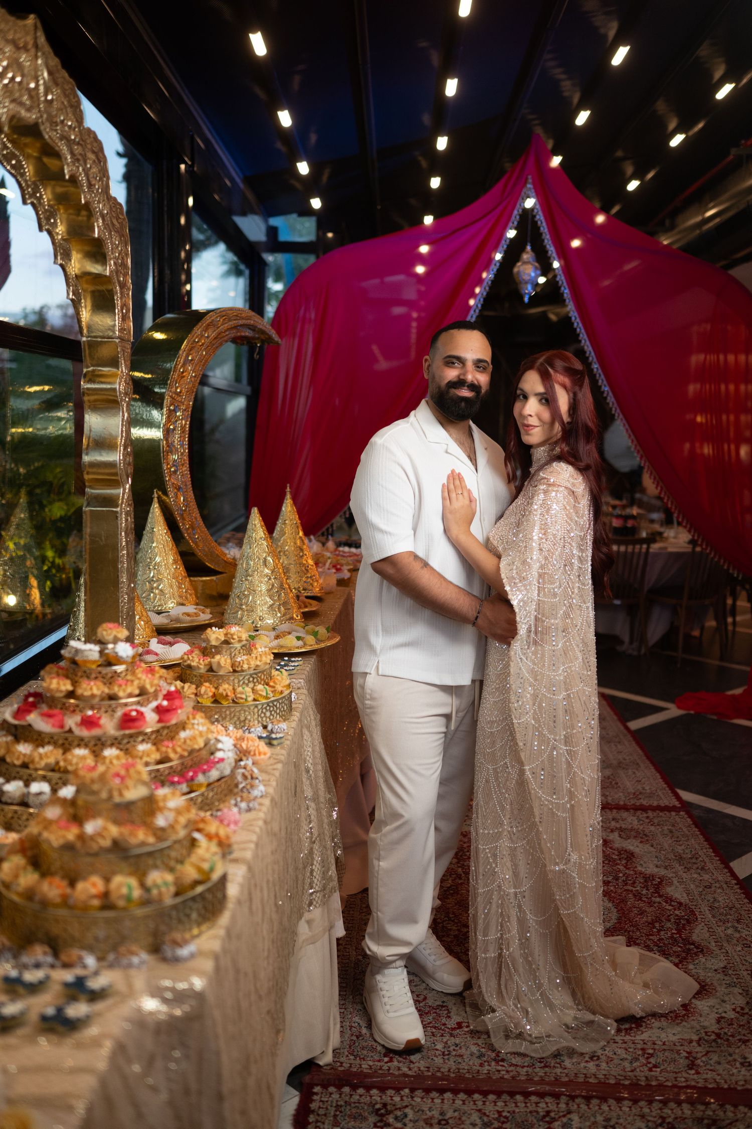 Couple in white attire posing by dessert table with ornate decor and red drape backdrop at elegant event.
