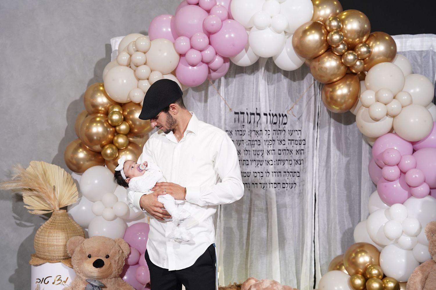 Baby celebration setup with pink, white and gold balloon arch and teddy bear decoration.