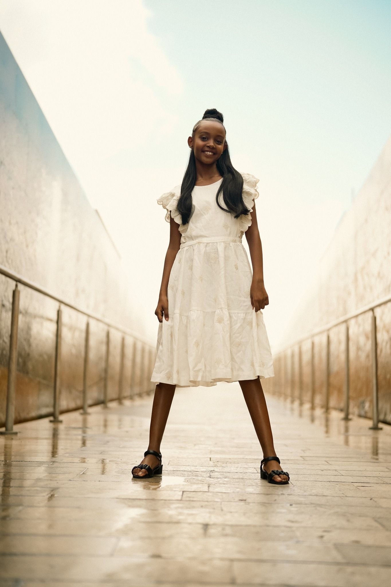 Person in white dress poses confidently on wet walkway between concrete walls.