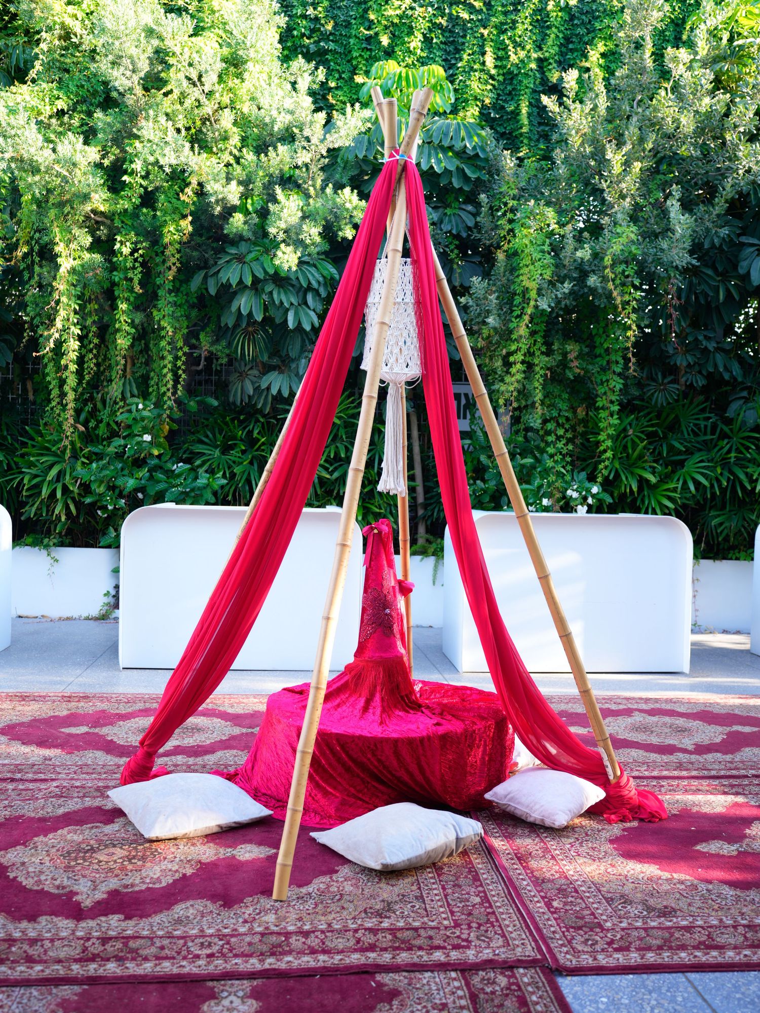 Outdoor wedding setup with red fabric draped teepee structures and white cushions on oriental rugs.