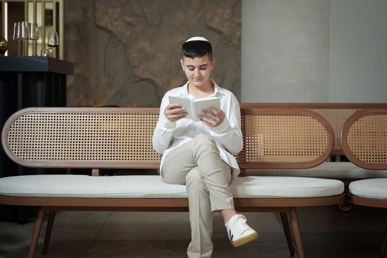 Person in beige sweater sits on woven cane bench while looking at mobile device.