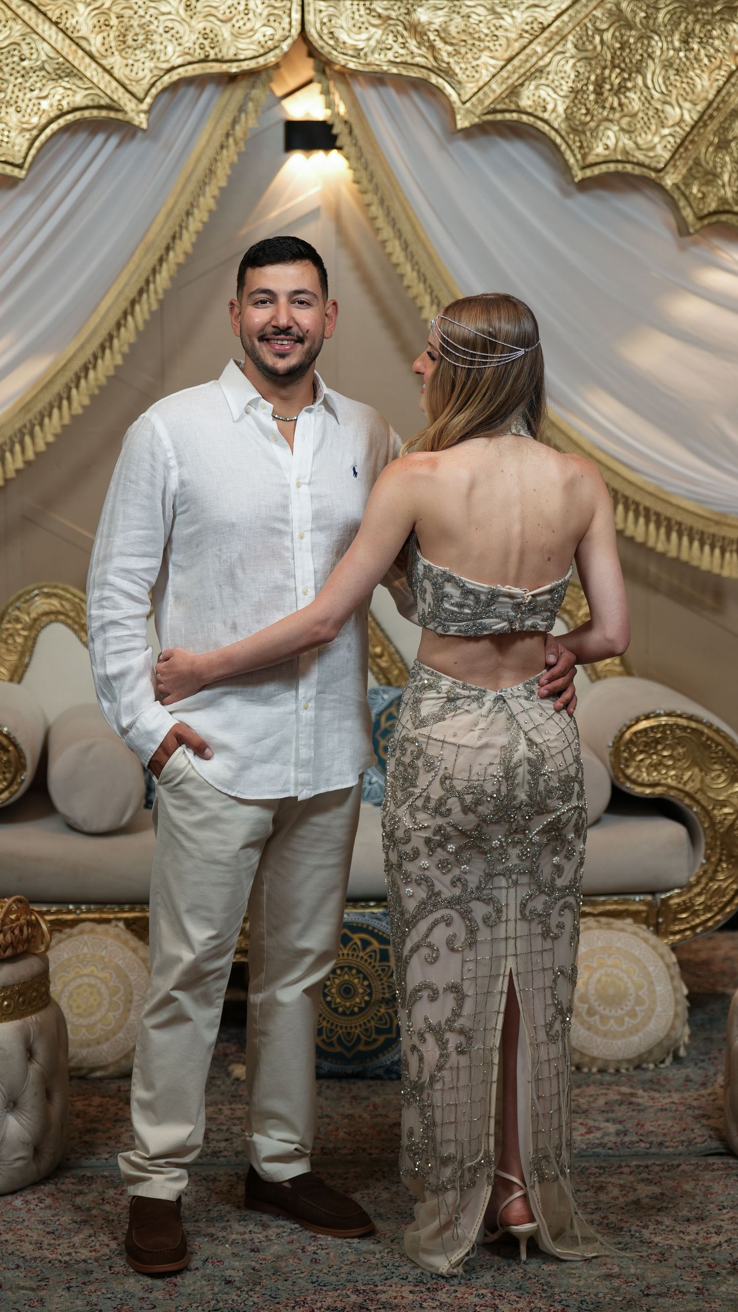 Elegant couple posing in ornate gold and white Moroccan-style decor, man in white shirt, woman in embellished gown.