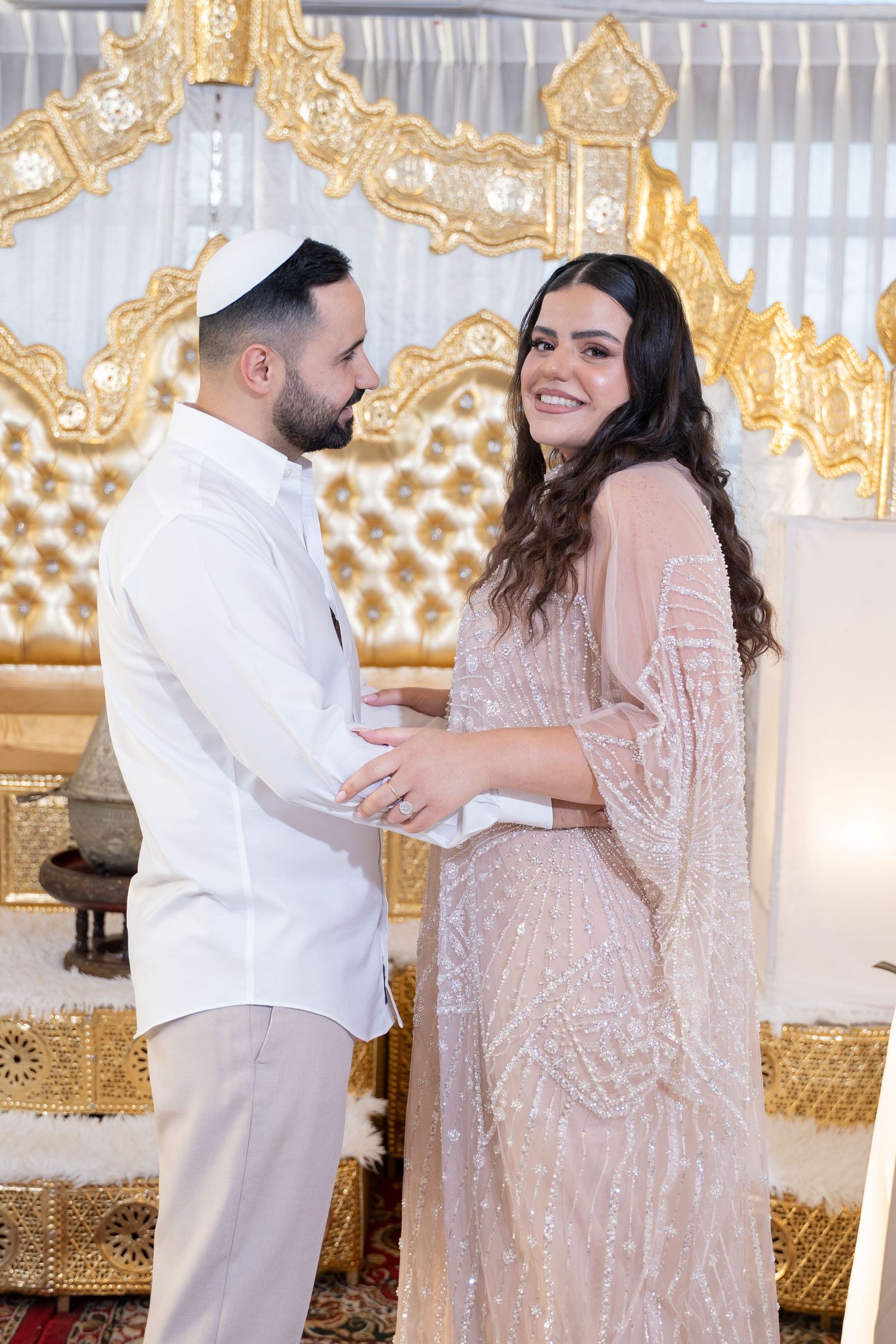 Couple shares intimate moment in front of luxurious gold decor wearing formal white and sparkly dress.