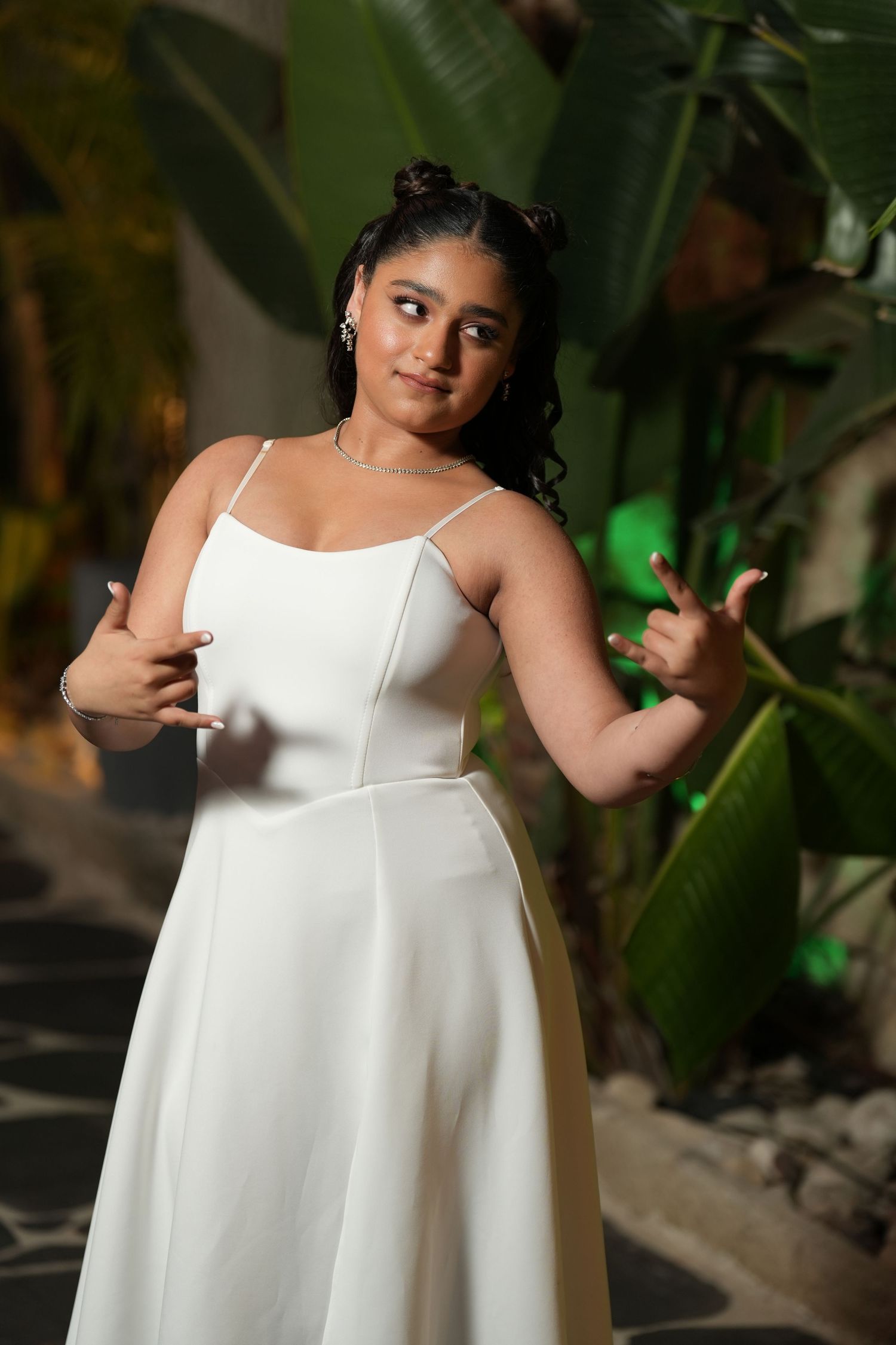 Young woman in white spaghetti strap dress posing outdoors with tropical plants in background.