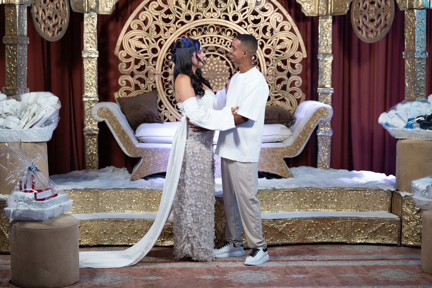 Couple in elegant attire stands before ornate gold throne backdrop at a lavish wedding ceremony.