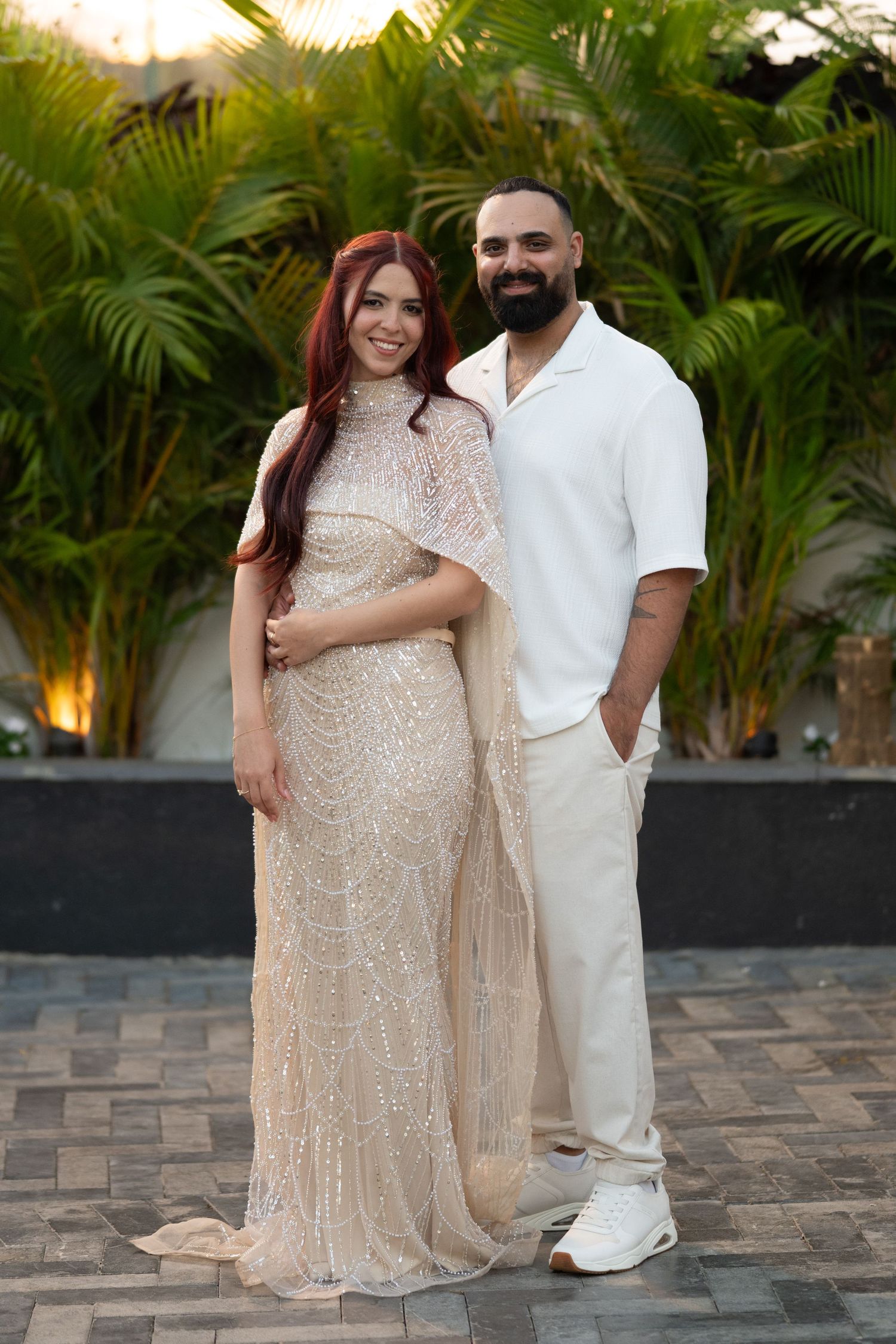 Couple posing outdoors in elegant white outfits, woman in sequined gown with red hair, man in casual white ensemble, tropical backdrop.