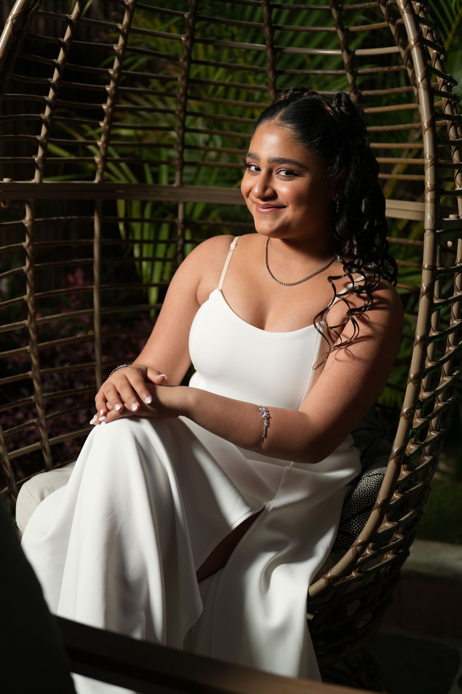 Young woman in white outfit sitting gracefully in a wicker egg chair, smiling softly with green foliage background.