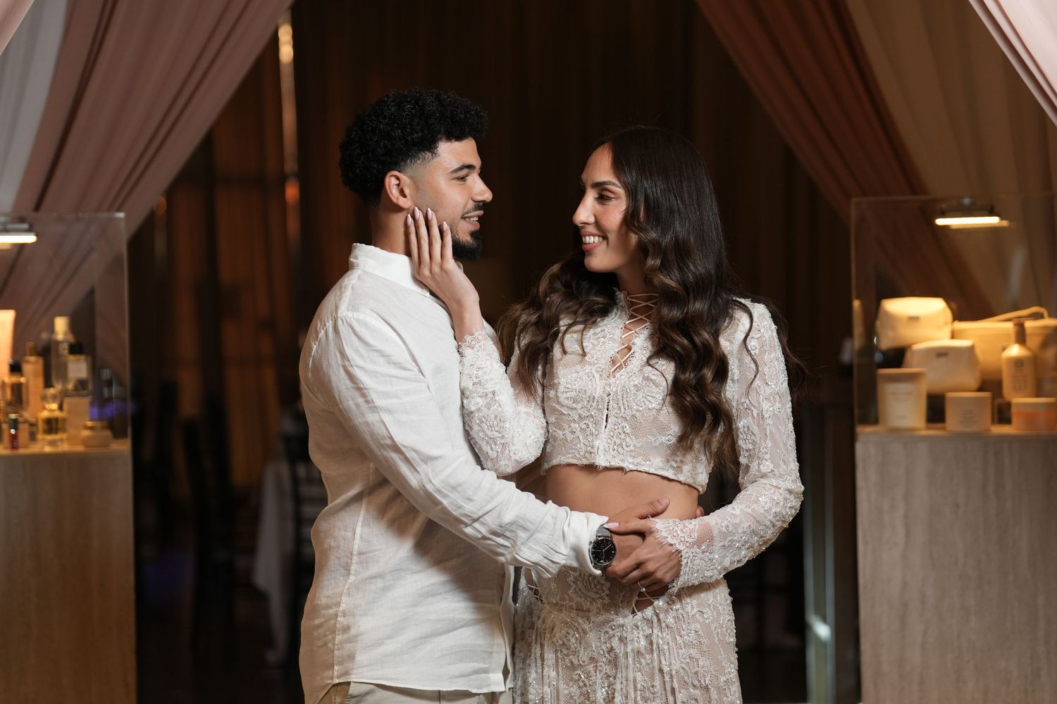 Couple in elegant white outfits sharing a romantic moment at an indoor event, woman touching man's face tenderly.
