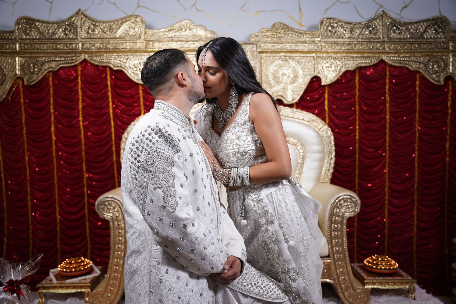 A romantic moment captured between a couple in matching white outfits against a red and gold backdrop.