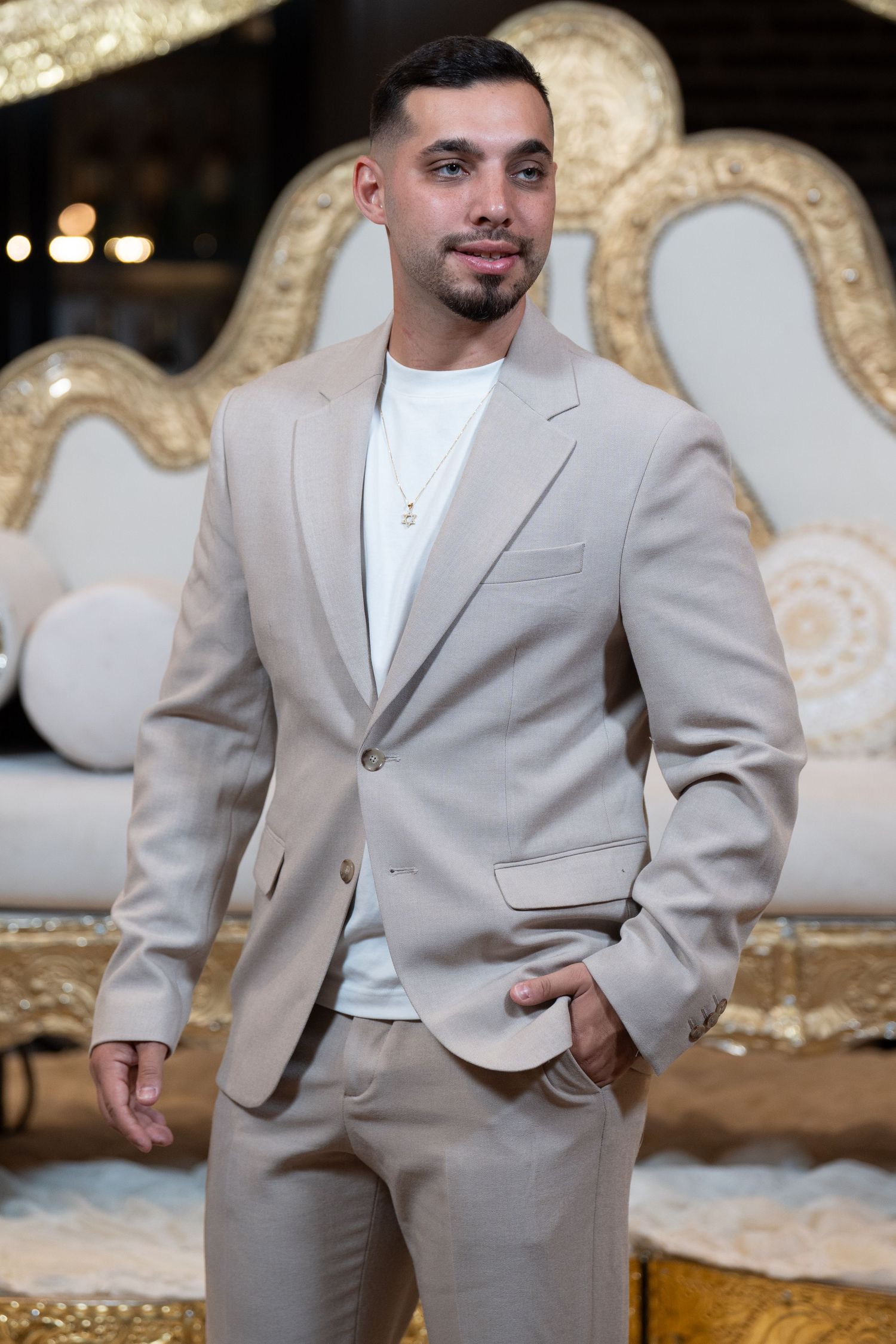 Man in beige suit and white shirt with gold necklace posing confidently in front of ornate white and gold decor.