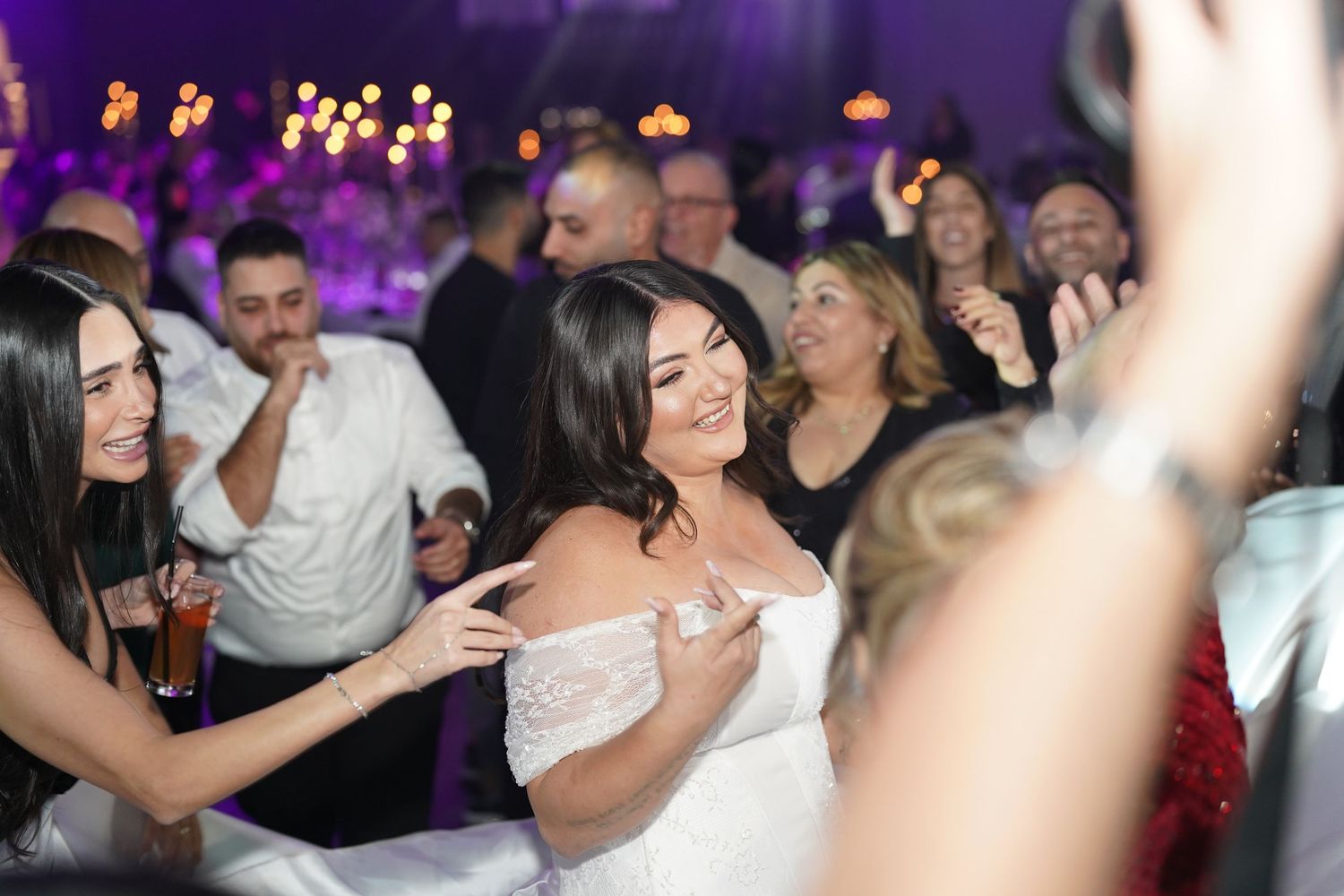 Bride in white off-shoulder gown laughing with guests on dance floor at elegant wedding reception.